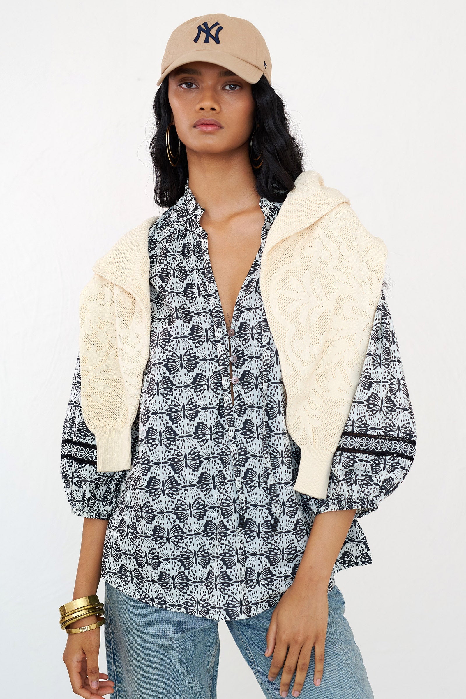 Front view of a woman standing. She is wearing the Lorna top in Flutter print styled with jeans, beige cardigan, and NY Yankees cap. The top features a V-neck with ruffle, tie-neck detail, voluminous sleeves, and a loose flowing body.