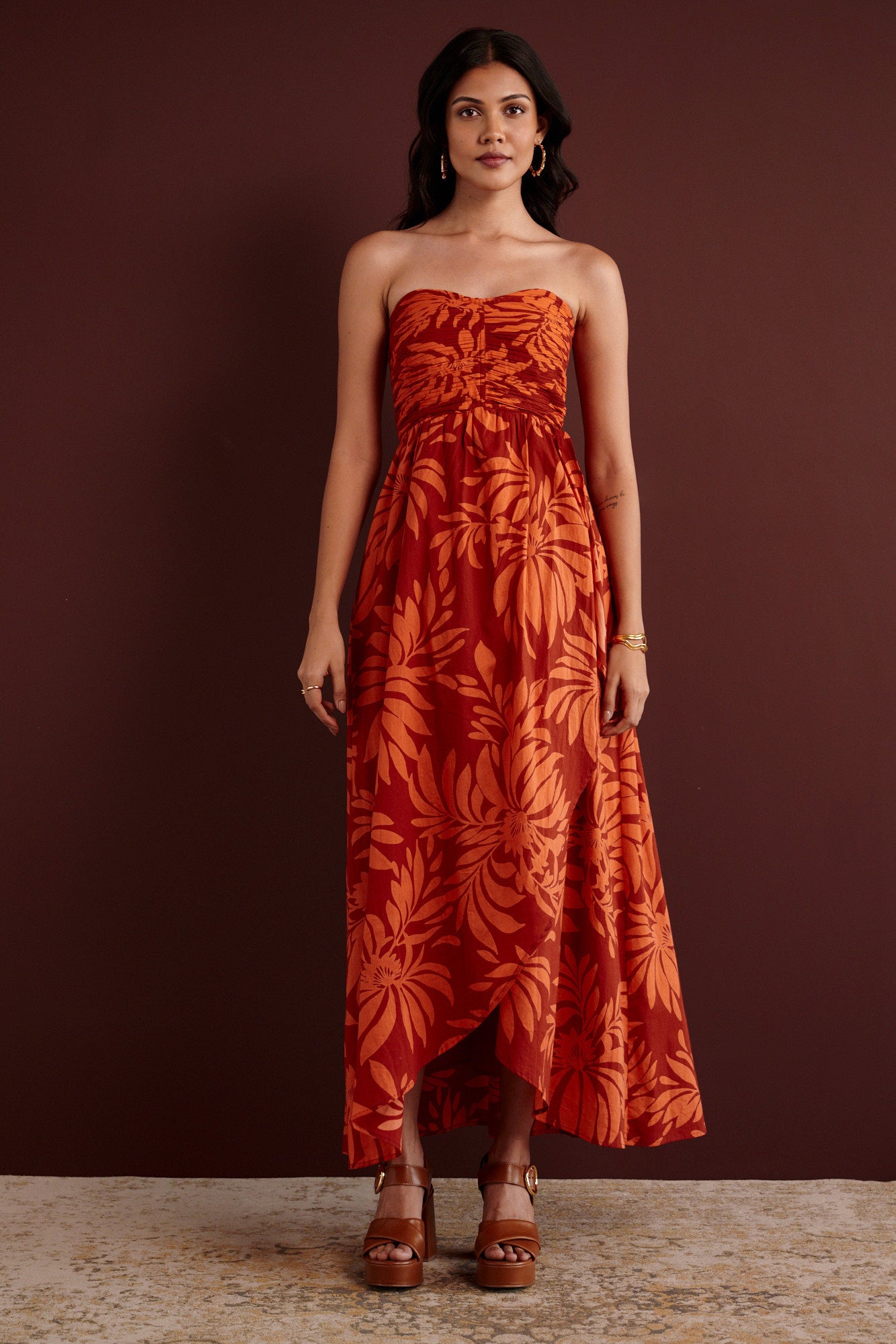 Front view of woman standing. She is wearing the Madison strapless maxi dress in Floral Frond print. It has a ruched bodice, cascading layered skirt, center back zipper, side-seam pockets, and bold Spice color inspired by pomegranate tree embroidery.