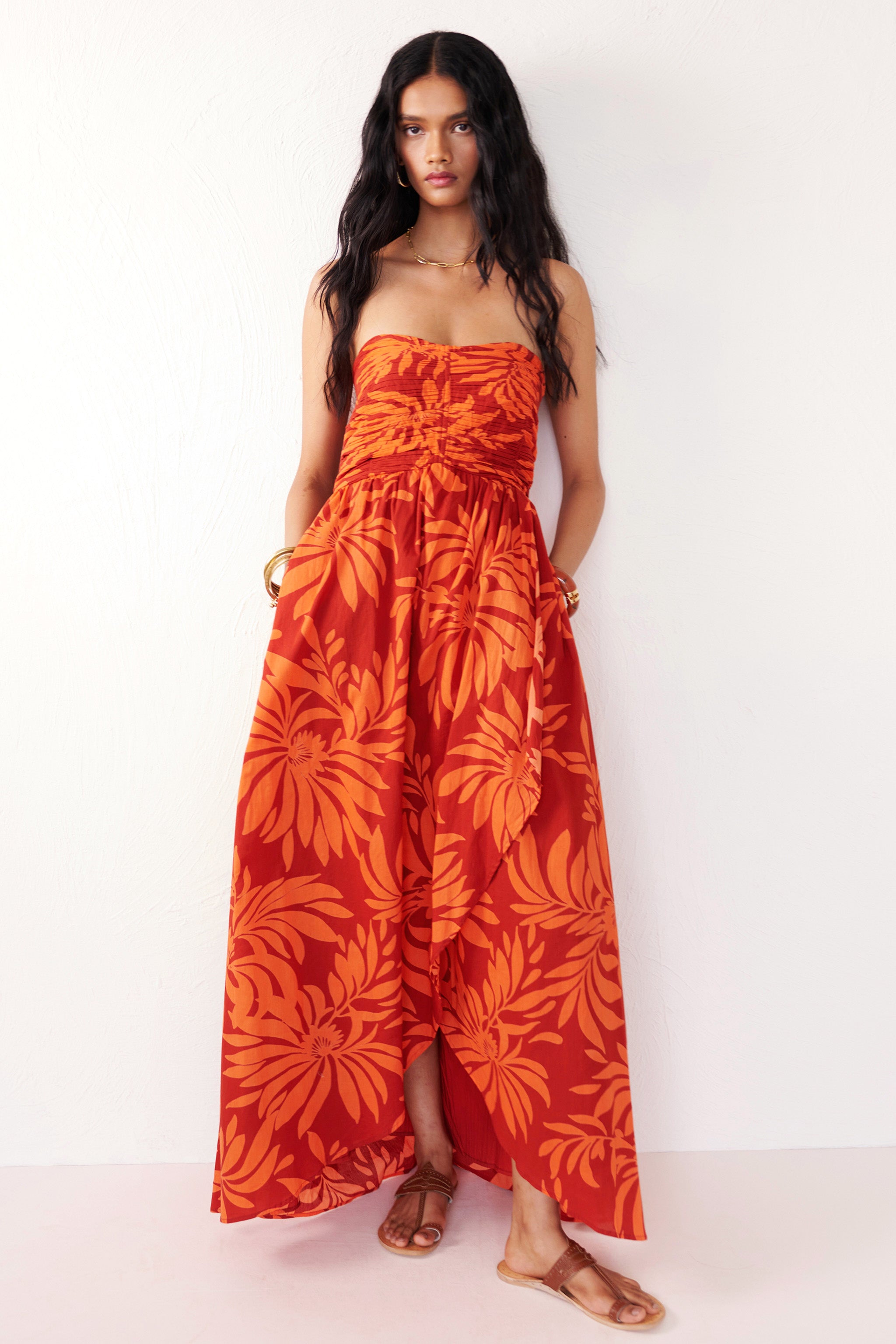 Front view of woman standing with hands in pocket. She is wearing the Madison strapless maxi dress in Floral Frond print, with ruched bodice, cascading layered skirt, center back zipper, side-seam pockets, and bold Spice color inspired by pomegranate tree embroidery.