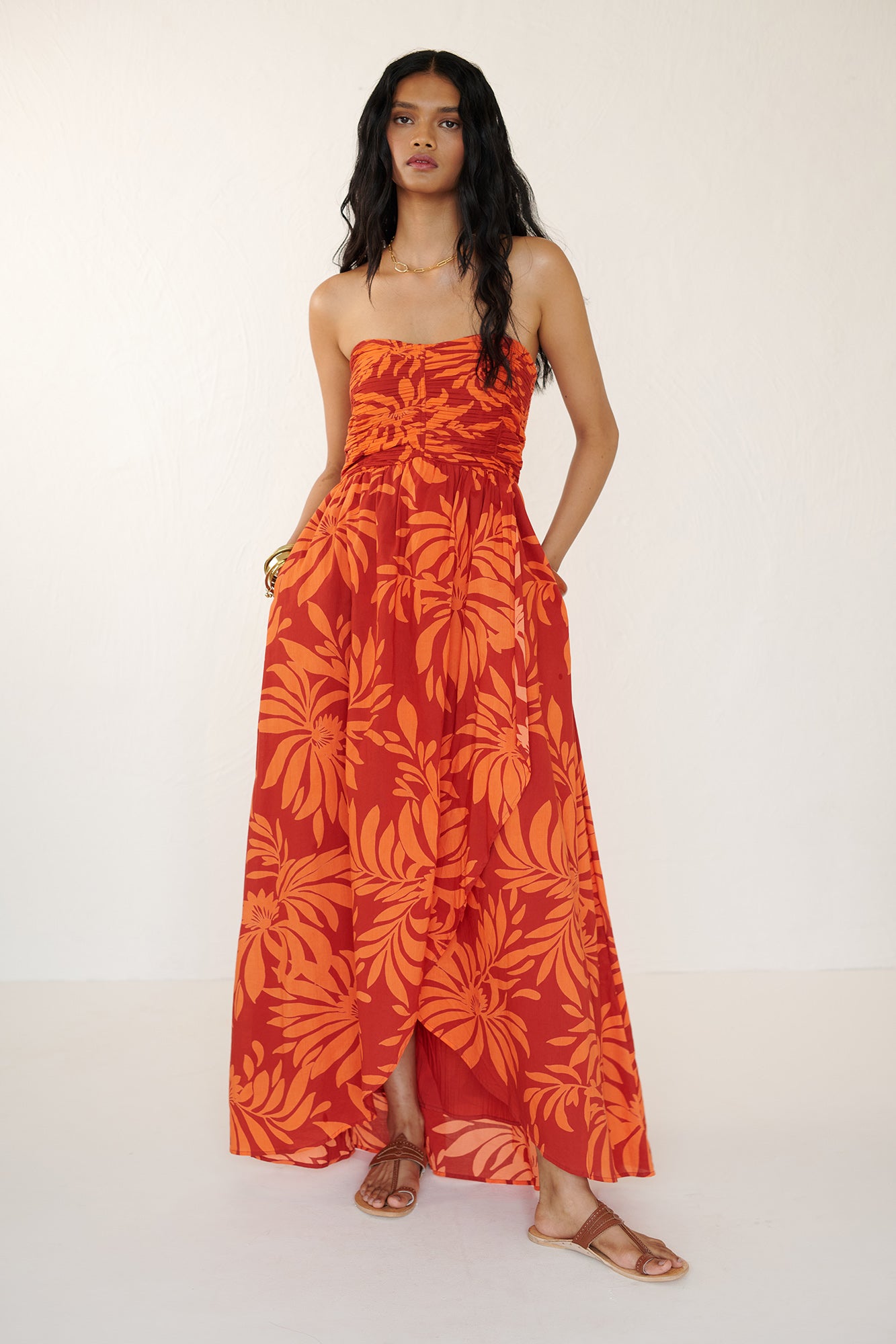 Front view of woman standing with both hands in pocket. She is wearing the Madison strapless maxi dress in Floral Frond print, with ruched bodice, cascading layered skirt, center back zipper, side-seam pockets, and bold Spice color inspired by pomegranate tree embroidery.
