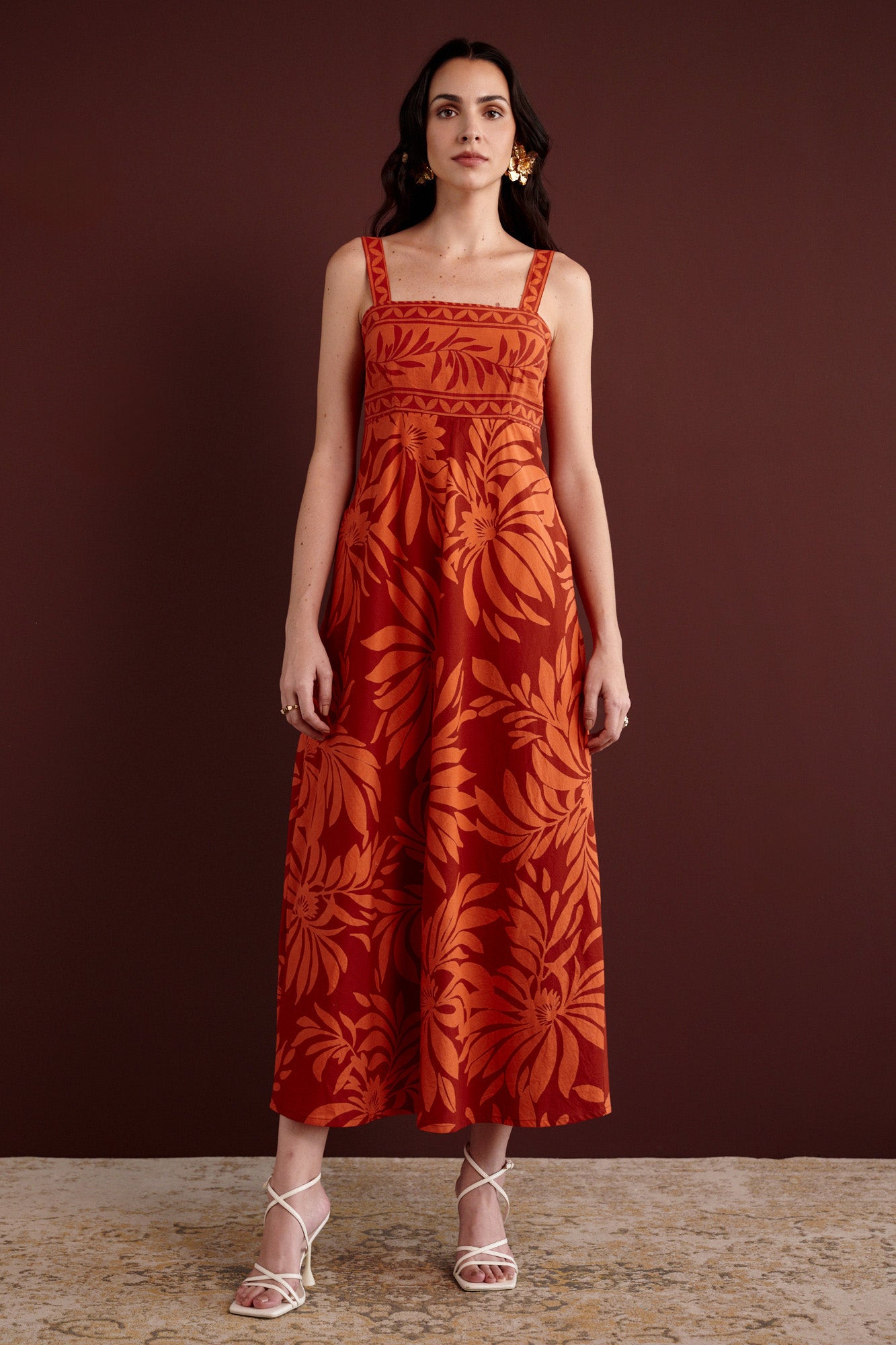 Front view of woman standing. She is wearing the Maria strappy midi dress in Floral Frond print. It is made from lightweight cotton voile, fully lined, with side-seam pockets, in a bold Spice color inspired by pomegranate tree embroidery.