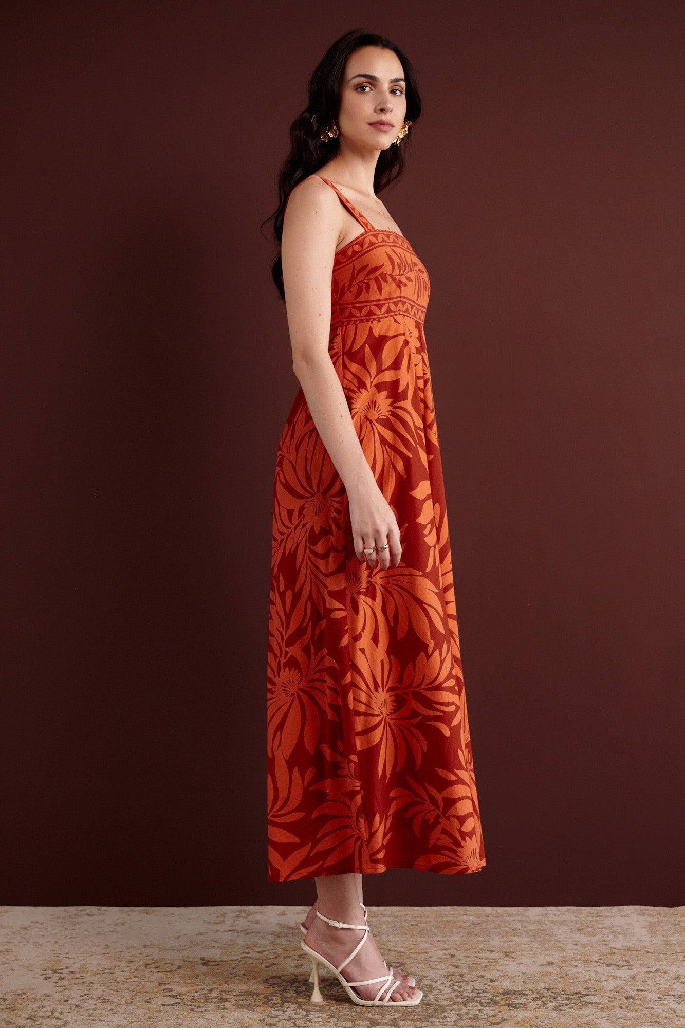 Side view of woman standing. She is wearing the Maria strappy midi dress in Floral Frond print. It is made from lightweight cotton voile, fully lined, with side-seam pockets, in a bold Spice color inspired by pomegranate tree embroidery.
