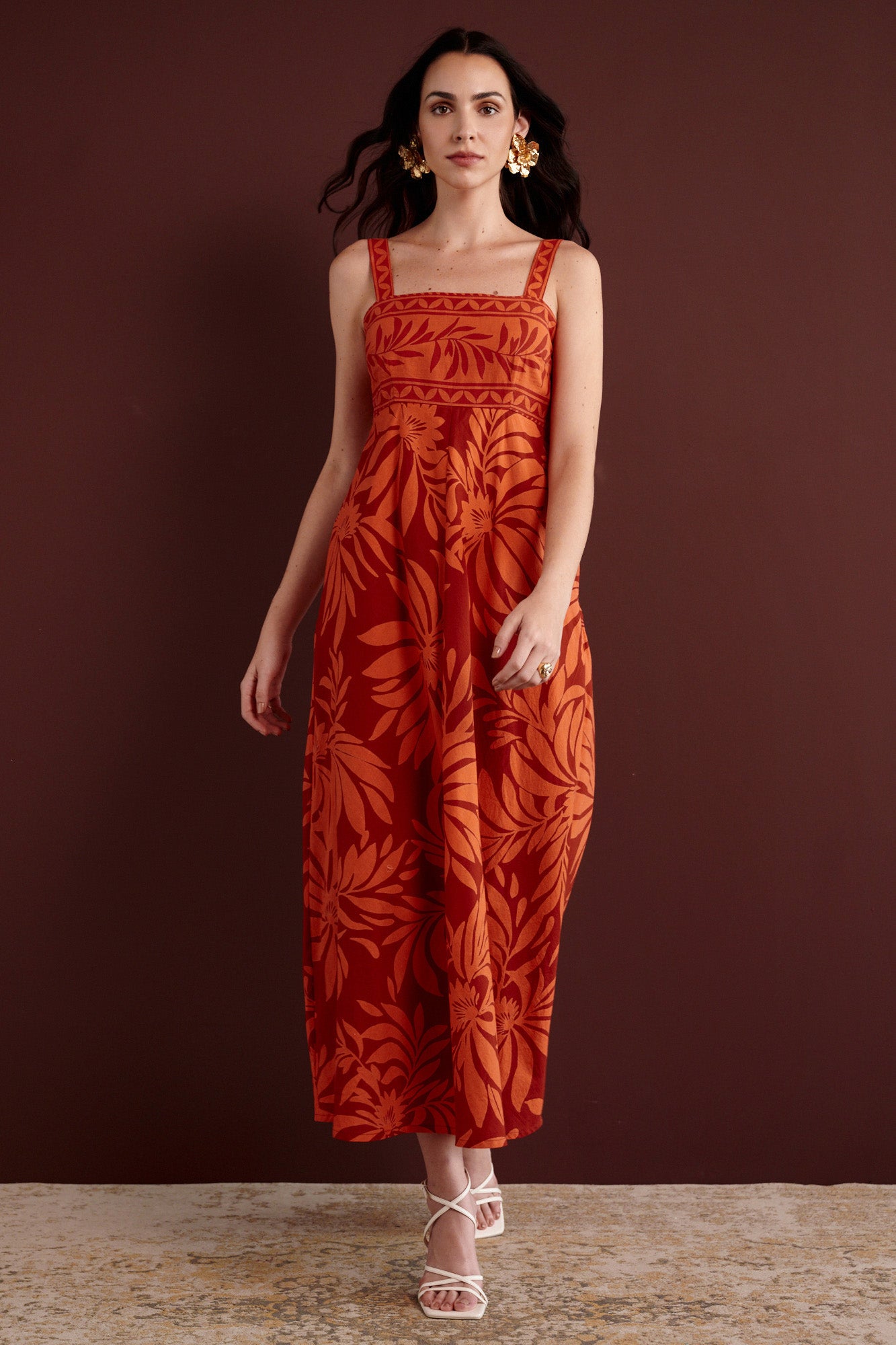 Front view of woman walking. She is wearing the Maria strappy midi dress in Floral Frond print. It is made from lightweight cotton voile, fully lined, with side-seam pockets, in a bold Spice color inspired by pomegranate tree embroidery.