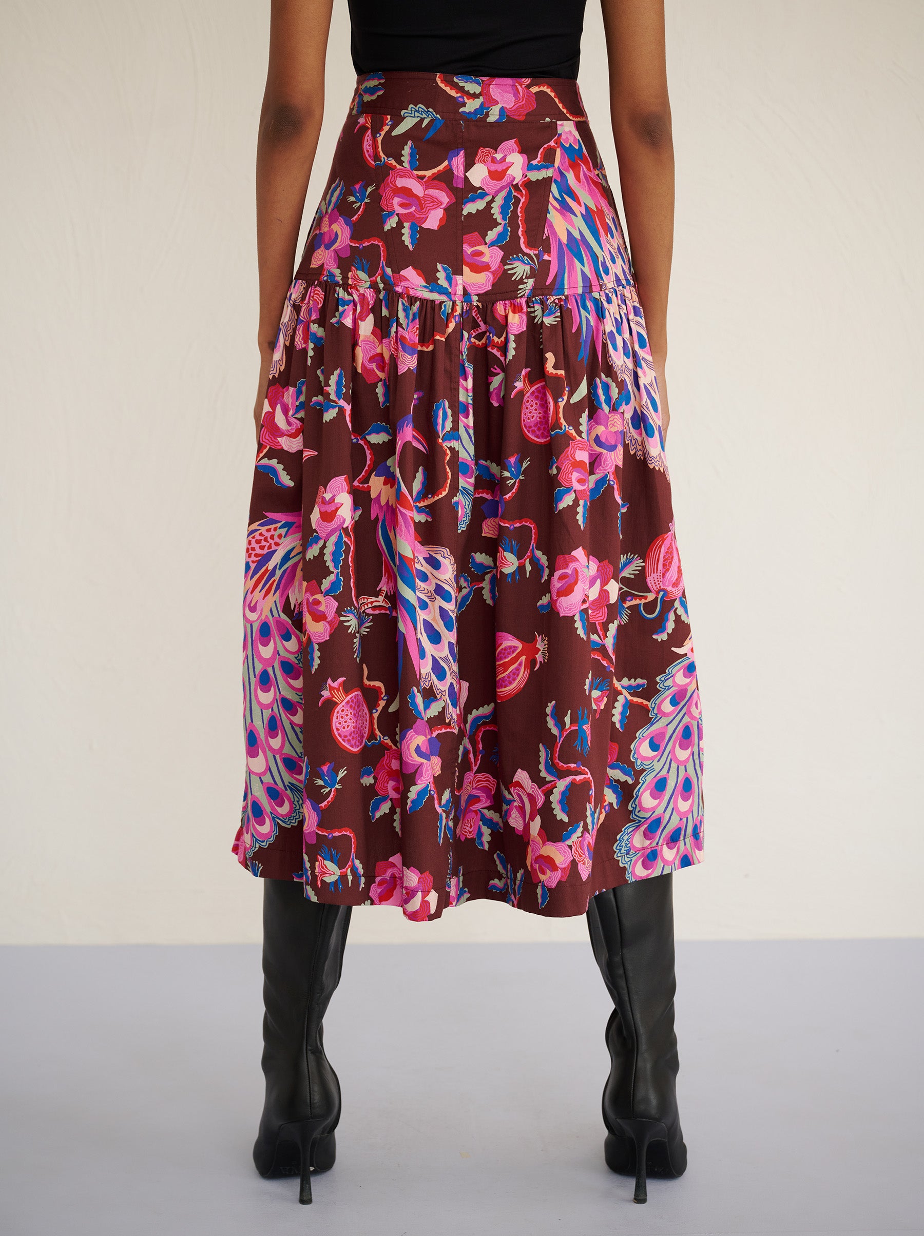 Back view close up of a model standing. She is wearing the Melodie skirt in cotton sateen with the Bukhara Peacock print in rum color. It is slim at the waist and gathered below for a flattering shape. The skirt is fully lined with side-seam pockets.
