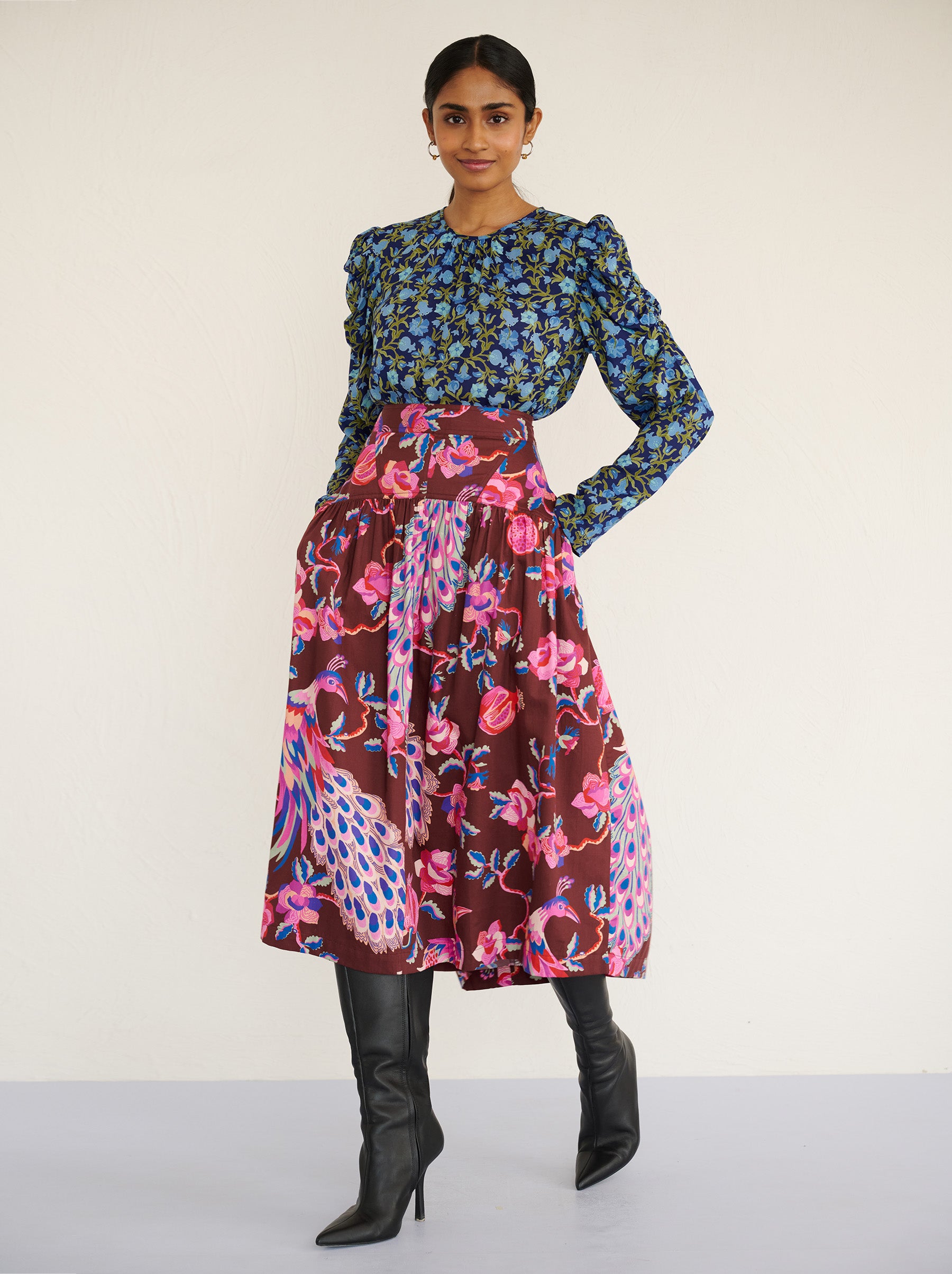 Front view of a model walking sideways. She is wearing the Melodie skirt in cotton sateen with the Bukhara Peacock print in rum color. It is slim at the waist and gathered below for a flattering shape. The skirt is fully lined with side-seam pockets. It is paired with Vincent top in Pomegrante Vine Lapis print with black boots.