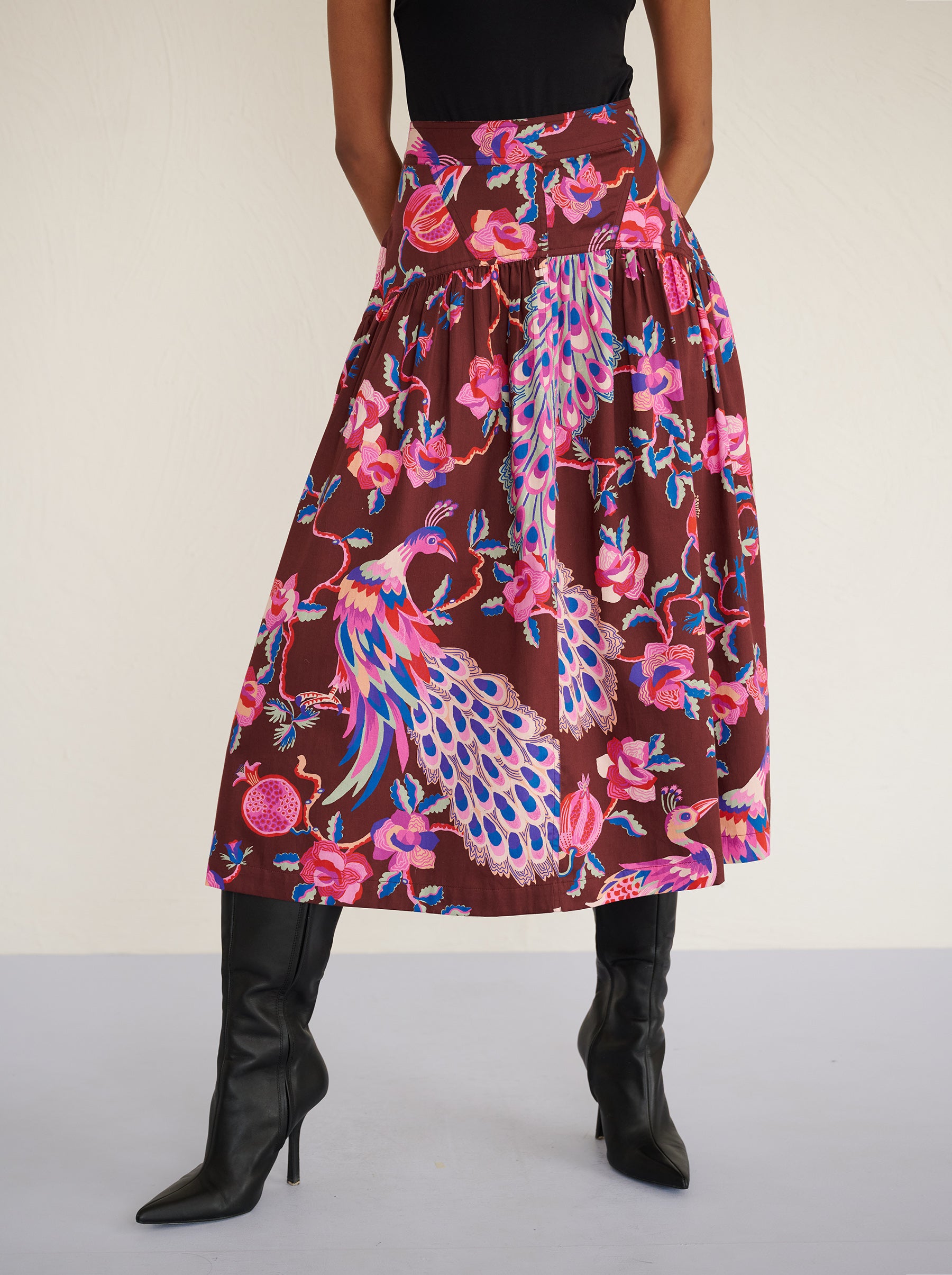 Front view close up of a model standing. She is wearing the Melodie skirt in cotton sateen with the Bukhara Peacock print in rum color. It is slim at the waist and gathered below for a flattering shape. The skirt is fully lined with side-seam pockets.