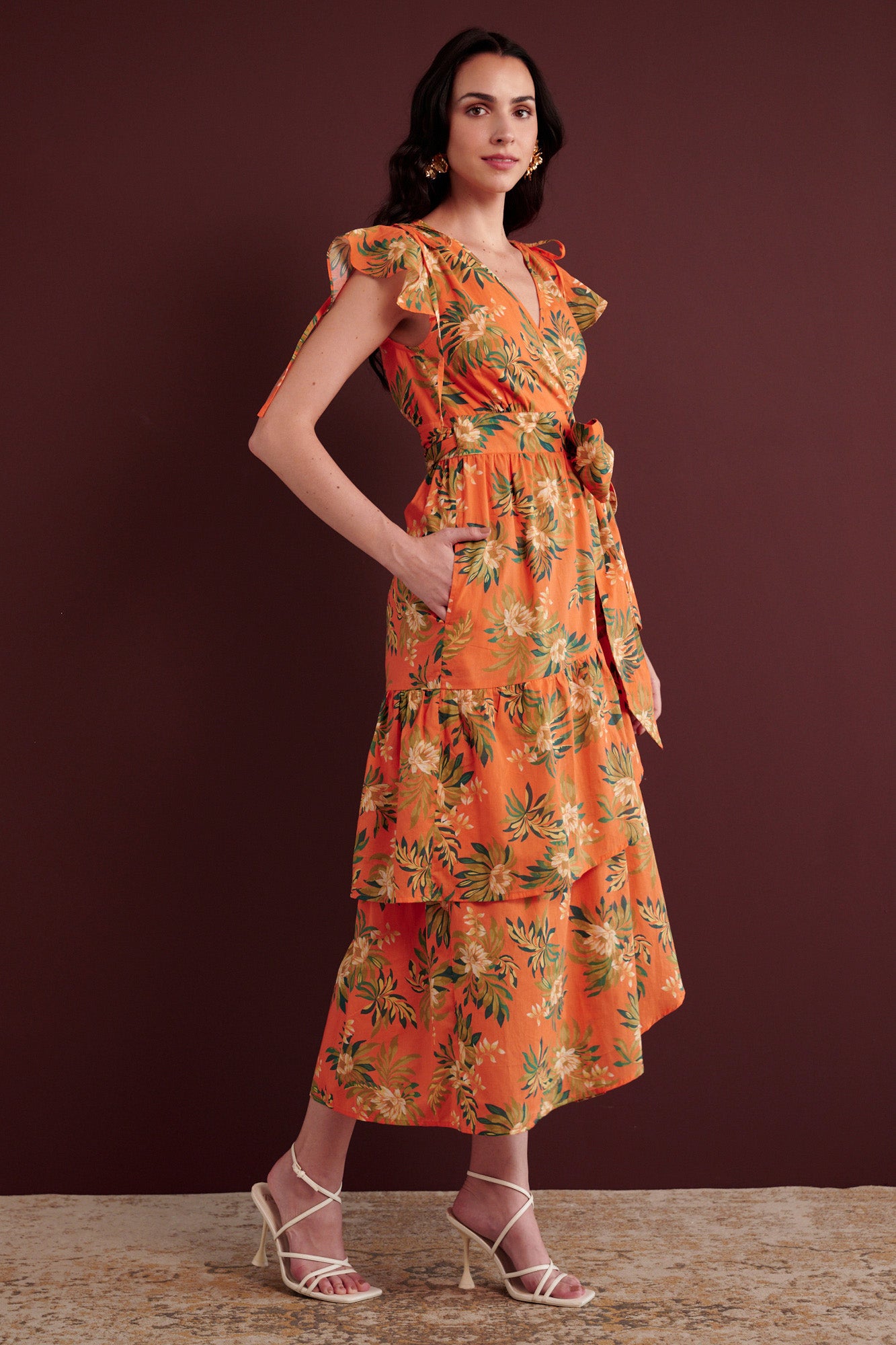Side view of woman standing with her hand in pocket. She is wearing the Mercy wrap dress in Artichoke Flower print. It has asymmetrical tiers, adjustable shoulder drawstrings, breezy cotton voile fabric, and side-seam pockets, inspired by 15th-century embroidered tapestry motifs.