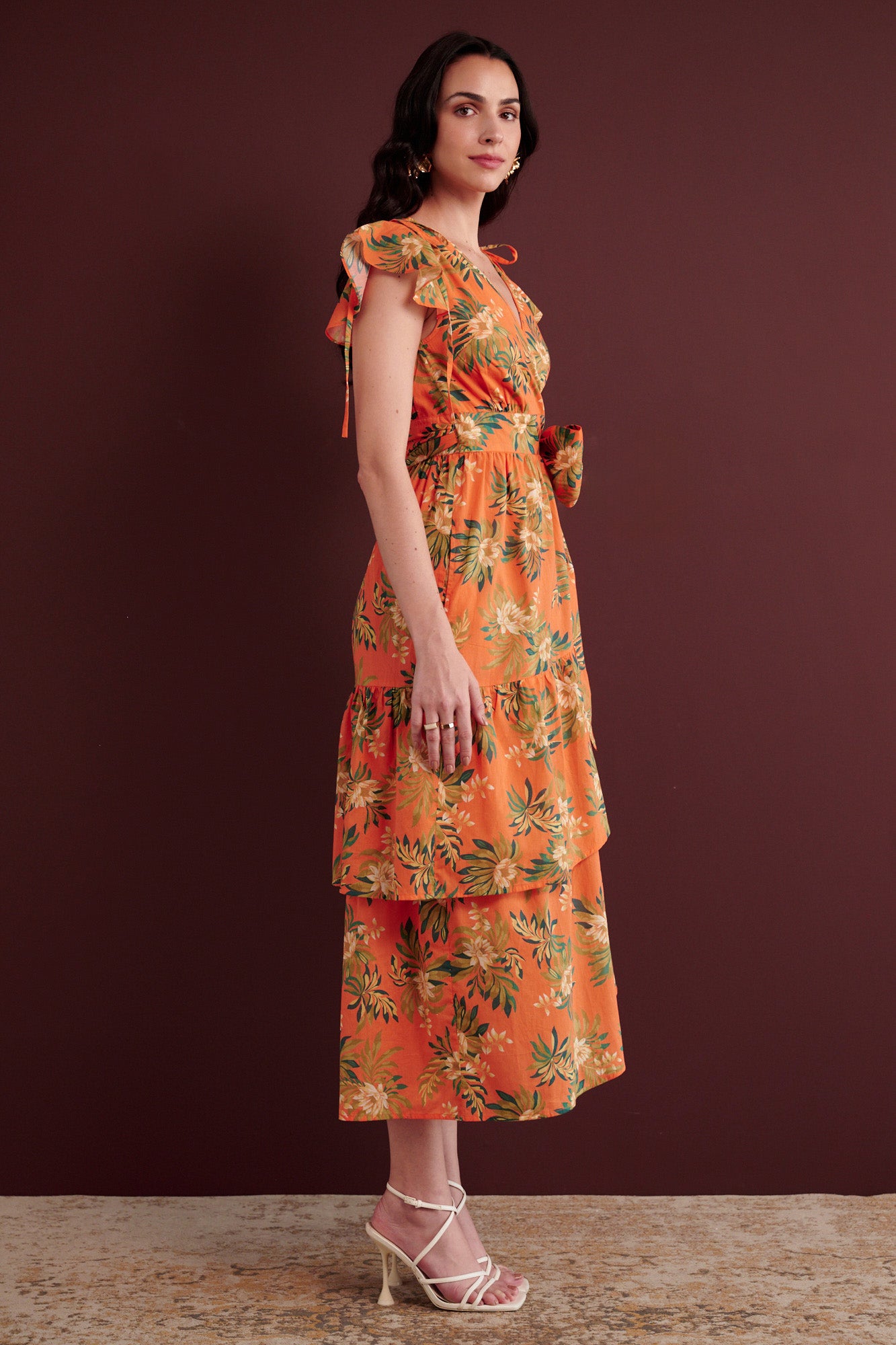 Side view of woman standing. She is wearing the Mercy wrap dress in Artichoke Flower print. It has asymmetrical tiers, adjustable shoulder drawstrings, breezy cotton voile fabric, and side-seam pockets, inspired by 15th-century embroidered tapestry motifs.