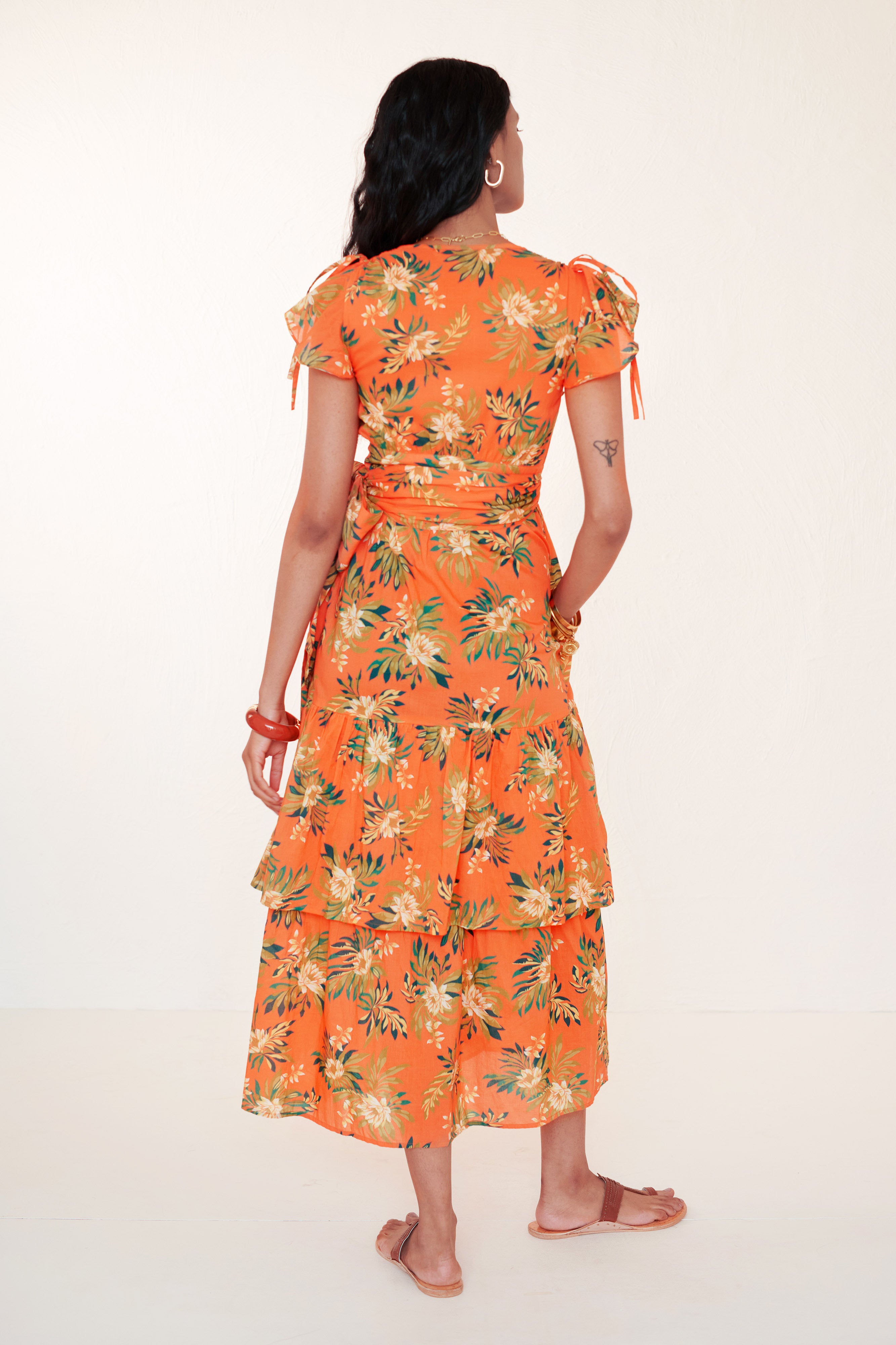 back view of woman standing with right hand in pocket. She is wearing the Mercy wrap dress in Artichoke Flower print. It has asymmetrical tiers, adjustable shoulder drawstrings, breezy cotton voile fabric, and side-seam pockets, inspired by 15th-century embroidered tapestry motifs.