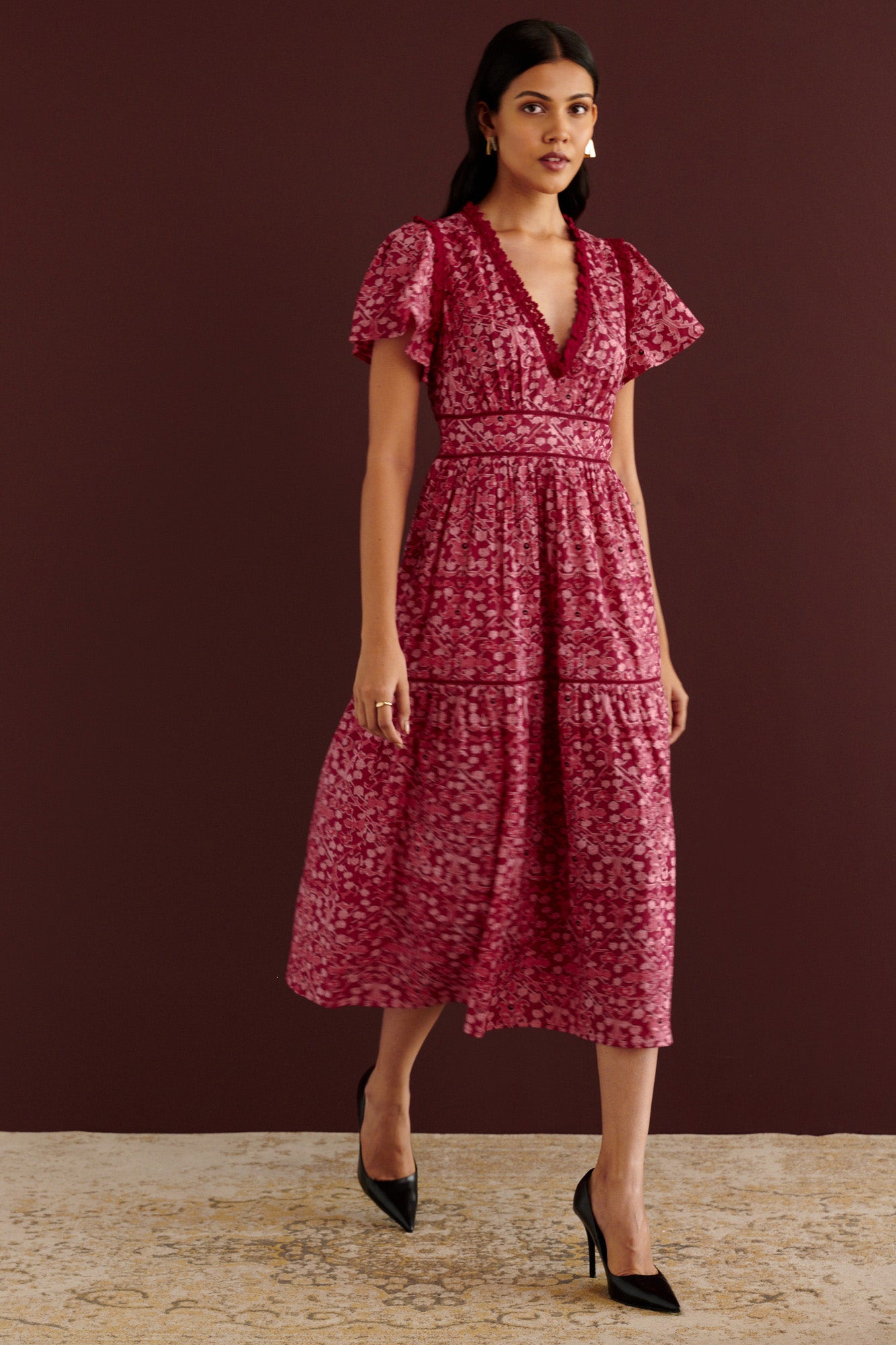 Front view of woman walking sideways. She is wearing the Naxos midi dress in Ottoman print, with plunging V-neck trimmed in red lace, lace insets at waist and skirt, flutter sleeves, smooth cotton cambric, fully lined construction, and side-seam pockets.