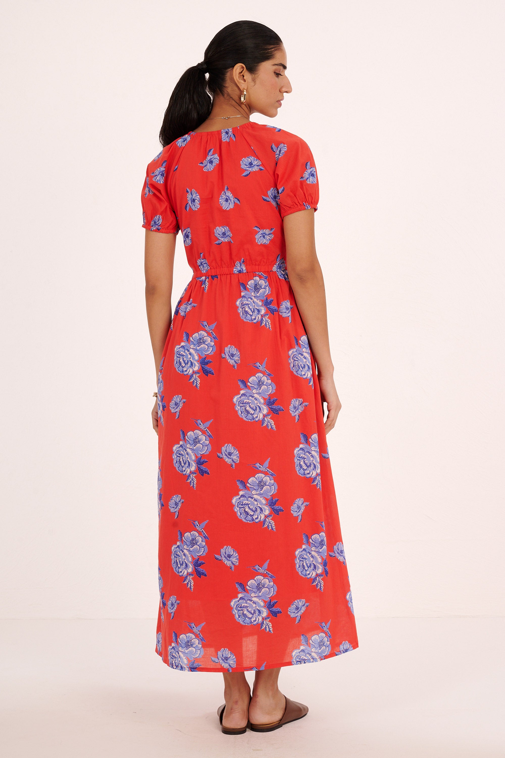 Back view of woman standing. She is  wearing the Norma dress in the Eliza Rose print, featuring a drawstring neckline, princess-seam waist, elastic sleeves, and floral motifs with blue roses and hummingbirds.