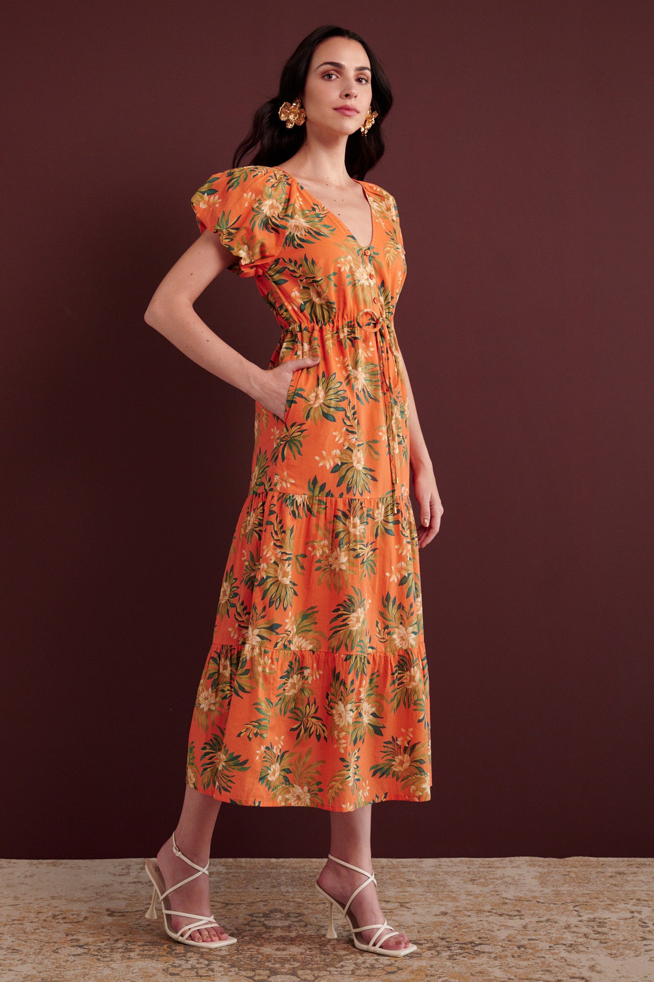 Side view of woman standing with her right hand in pocket. She is wearing the Poppy dress in Artichoke Flower print. It has a raglan blouson sleeves, front button placket, drawstring waist, flowing skirt, cap sleeves, fully lined construction, and side-seam pockets.