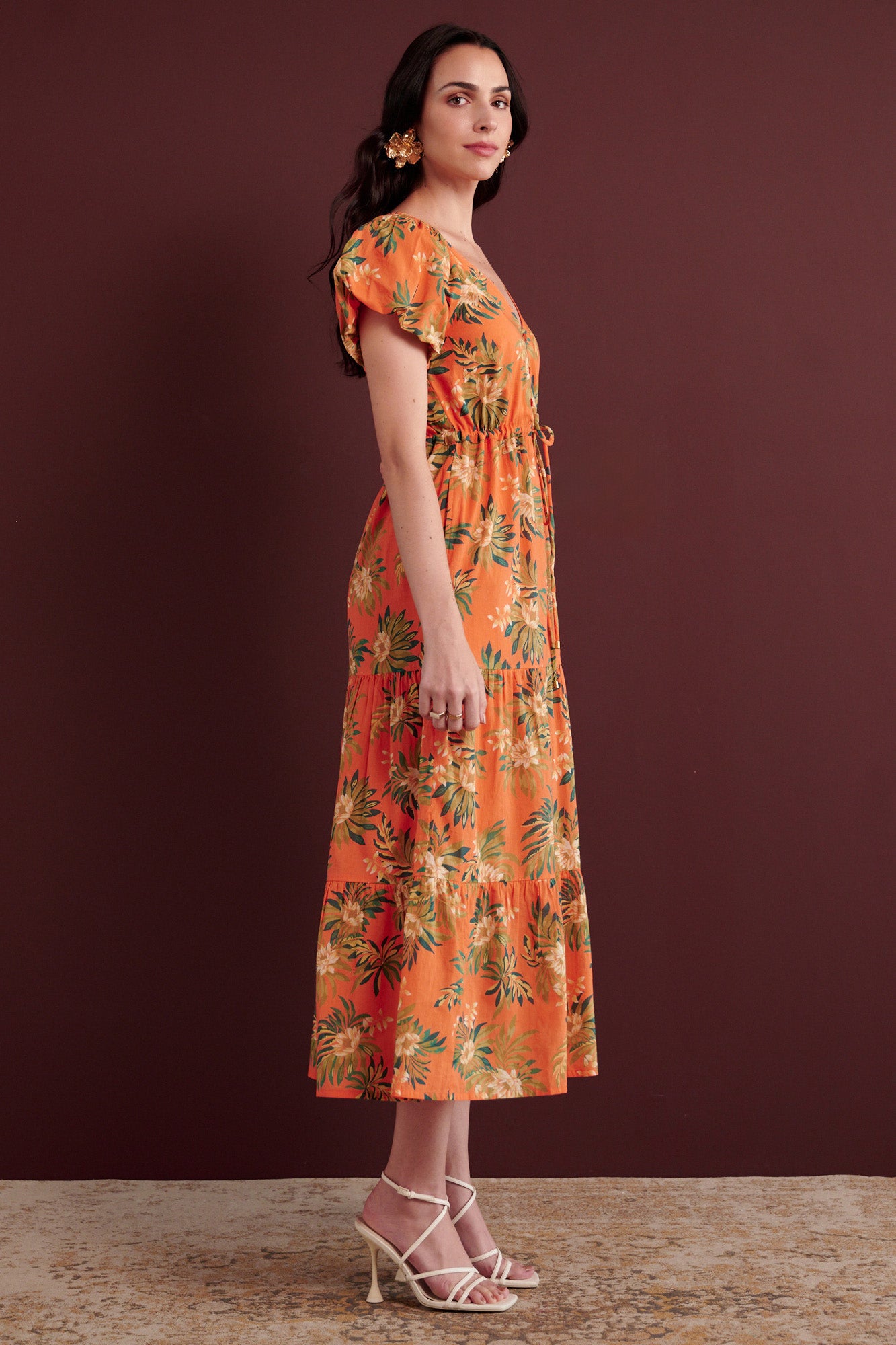 Side view of woman standing. She is wearing the Poppy dress in Artichoke Flower print. It has a raglan blouson sleeves, front button placket, drawstring waist, flowing skirt, cap sleeves, fully lined construction, and side-seam pockets.