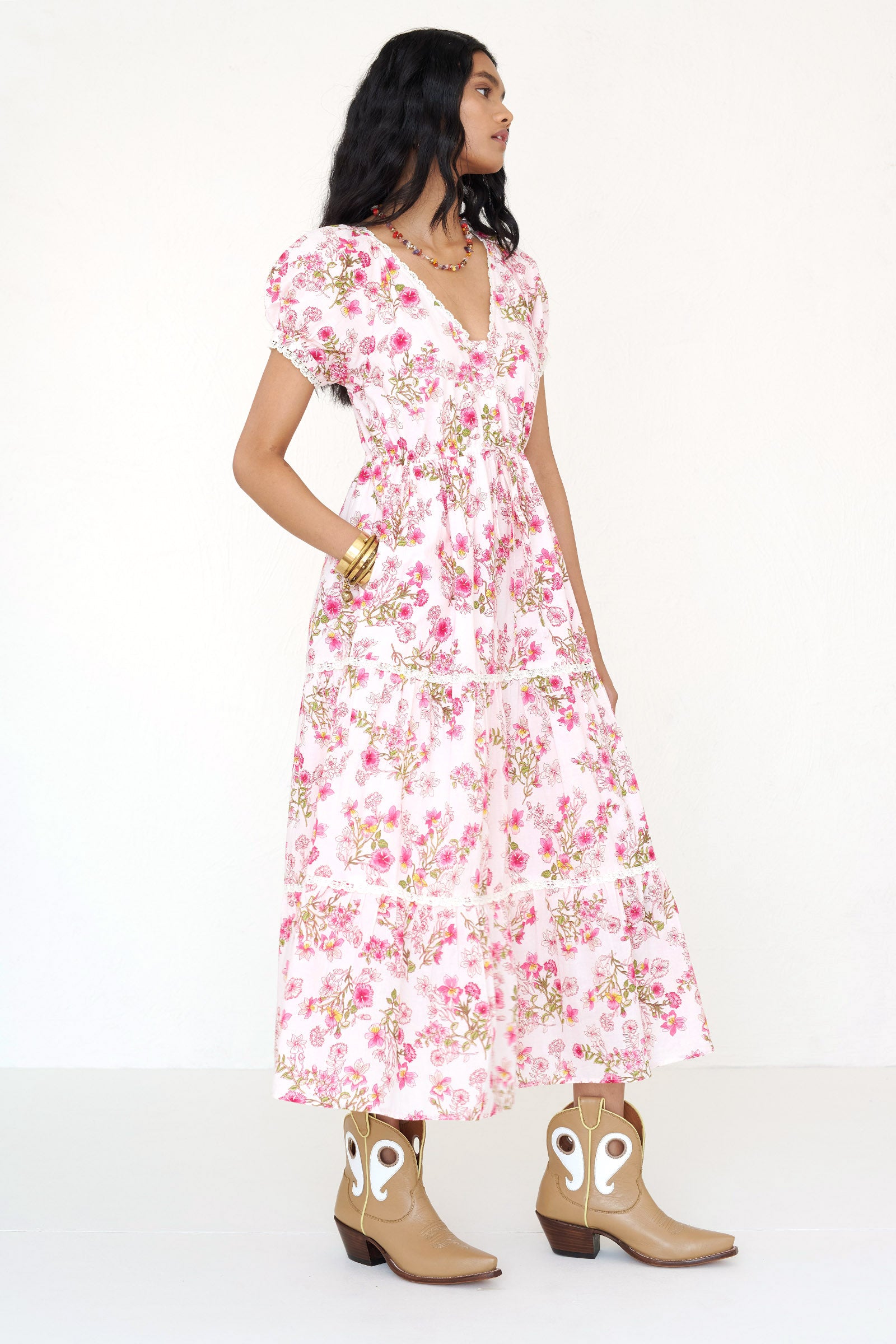Side view of a woman standing wearing the Poppy dress in Burdock floral print with her hand in pocket.It features raglan blouson sleeves, front button placket, drawstring waist, lace-trimmed tiers, flowing skirt, and side-seam pockets. 