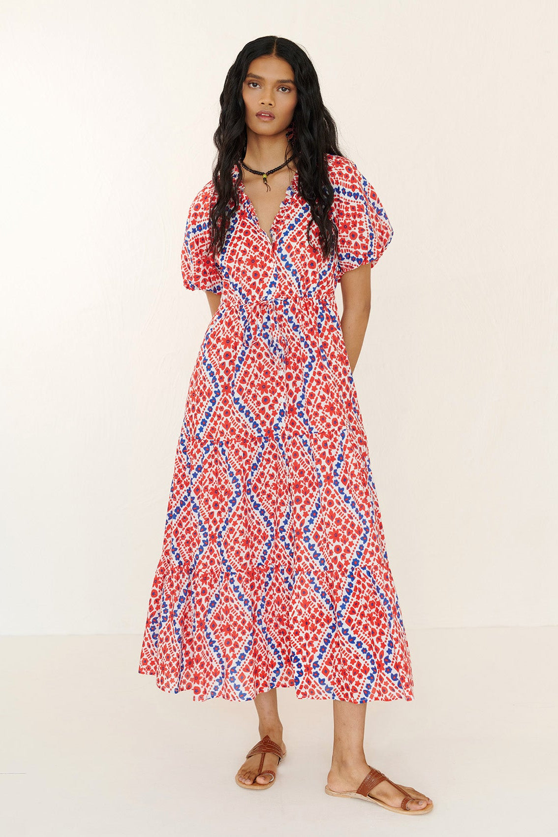 Front view of woman standing. She is wearing Poppy Dress in Athena Heart print poppy color. Its is a dress with floral and heart motifs, blouson sleeves, button front, drawstring waist, and flowing long skirt.