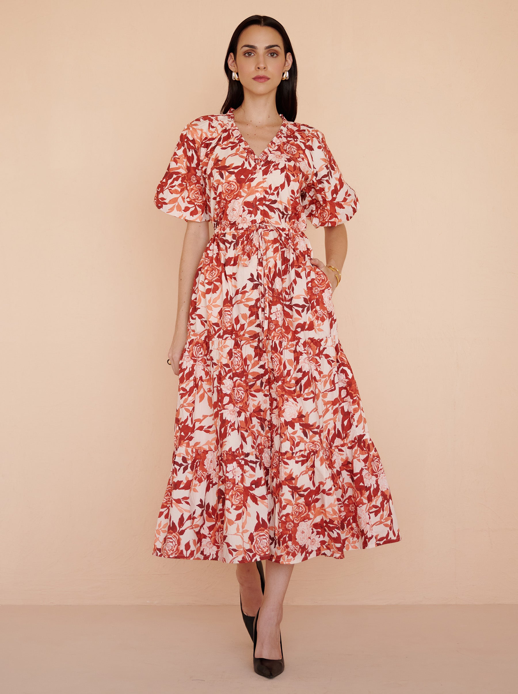 Front view of a model walking. She is wearing the Poppy dress in crisp cotton poplin with the Bibi Rose print in red-chocolate color flowers with cream base. It is featuring raglan blouson sleeves, a front button placket, drawstring waist, and a long flowing skirt. The dress is fully lined with side-seam pockets.