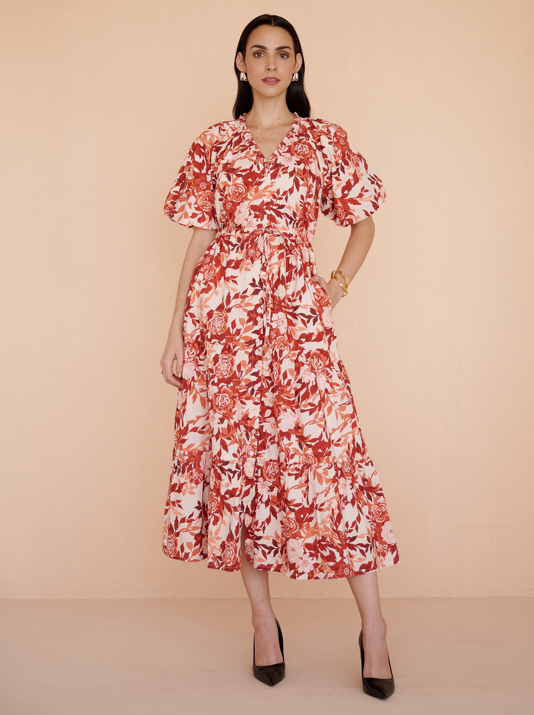 Front view of a model standing. She is wearing the Poppy dress in cotton poplin with the Bibi Rose print in red-chocolate color flowers with cream base.It is  featuring raglan blouson sleeves, a front button placket, drawstring waist, and a long flowing skirt. The dress is fully lined with side-seam pockets.