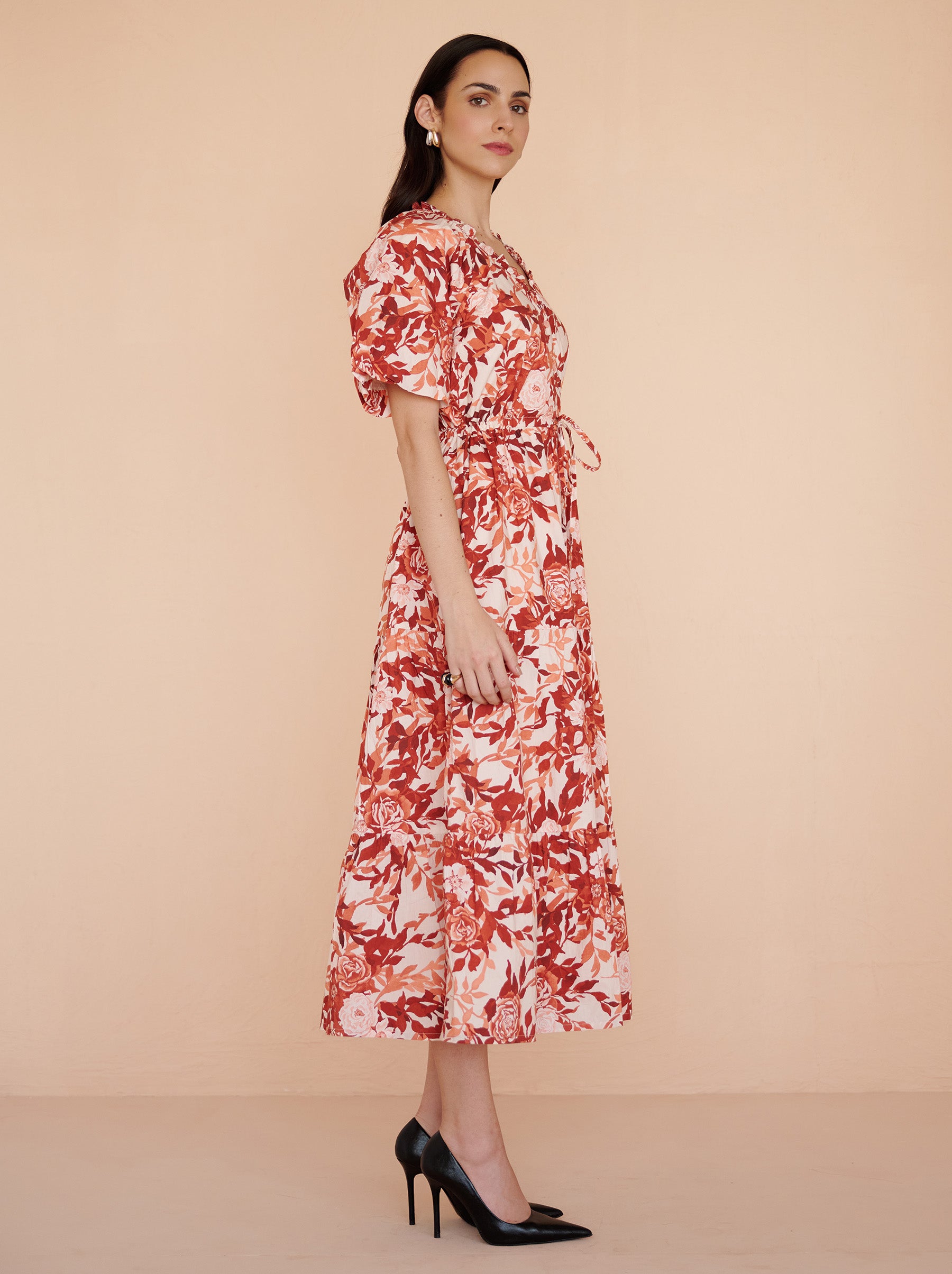Side view of a model standing. She is wearing the Poppy dress in crisp cotton poplin with the Bibi Rose print in red-chocolate color flowers with cream base. It is featuring raglan blouson sleeves, a front button placket, drawstring waist, and a long flowing skirt. The dress is fully lined with side-seam pockets.