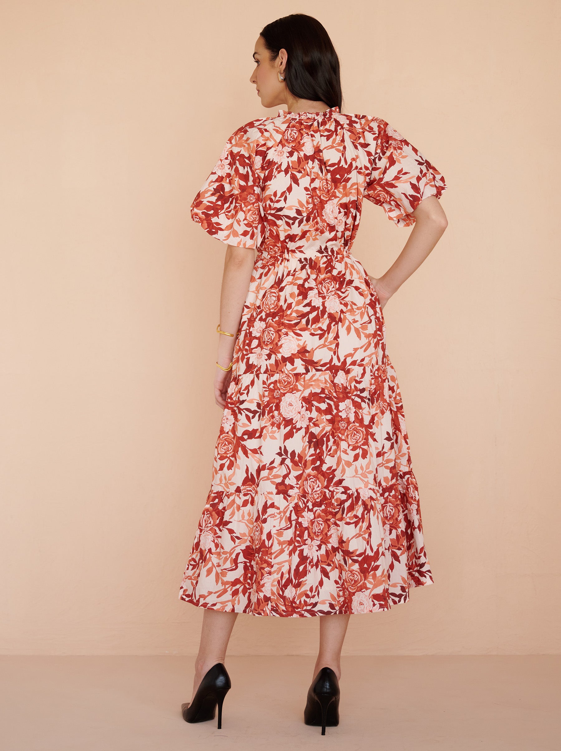 Back view of a model standing. She is wearing the Poppy dress in crisp cotton poplin with the Bibi Rose print in red-chocolate color flowers with cream base.It is  featuring raglan blouson sleeves, a front button placket, drawstring waist, and a long flowing skirt. The dress is fully lined with side-seam pockets.