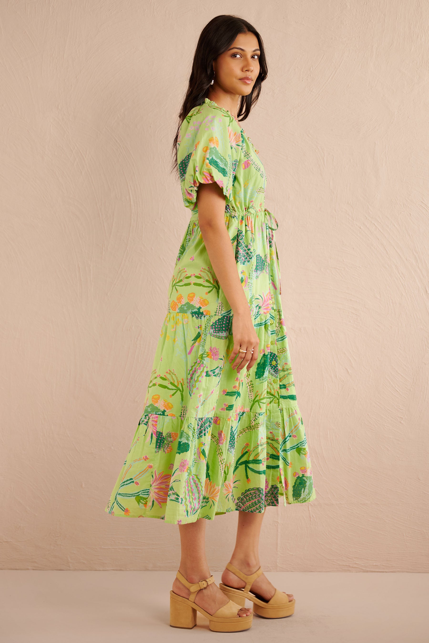 Side view of the Poppy dress in a green Calypso Cacti print with blouson sleeves, button front, drawstring waist, and a flowing tiered skirt featuring desert blooms, birds, and butterflies.