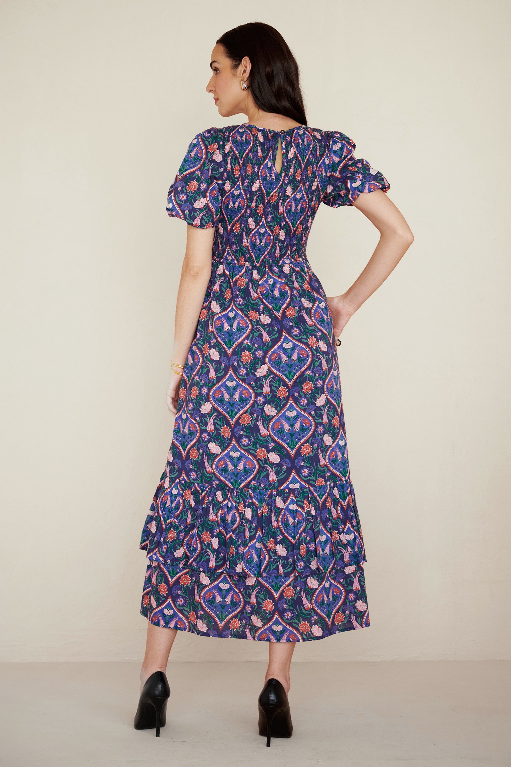 Back view of woman wearing a Quant dress in Persian Tile print in Lapis color with her right hand in pocket. It is a midi dress with smocked bodice, puff sleeves, and tiered skirt, featuring a floral tile print inspired by Uzbek design.