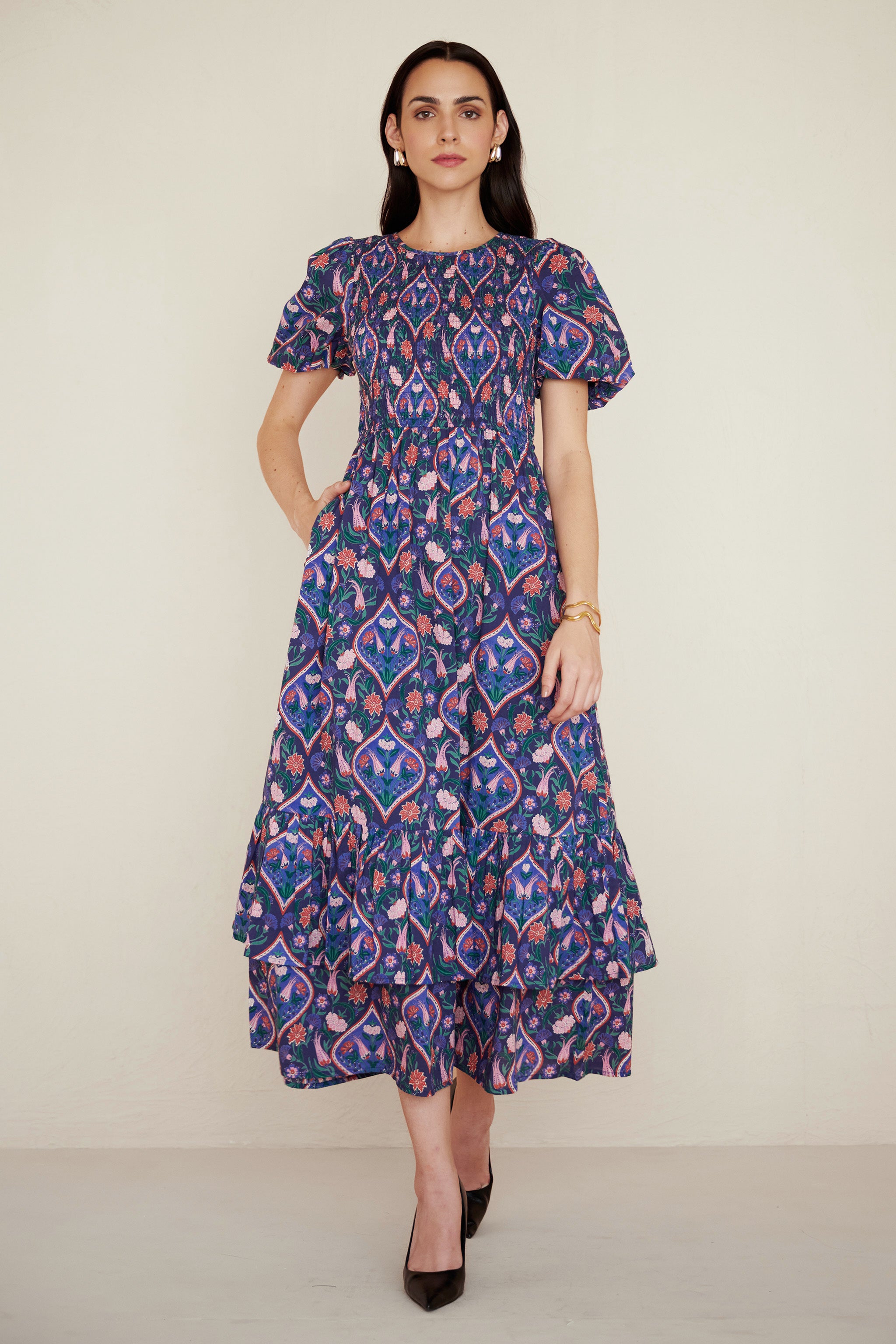 Front view of woman wearing a Quant dress in Persian Tile print in Lapis color with her right hand in pocket. It is a midi dress with smocked bodice, puff sleeves, and tiered skirt, featuring a floral tile print inspired by Uzbek design.