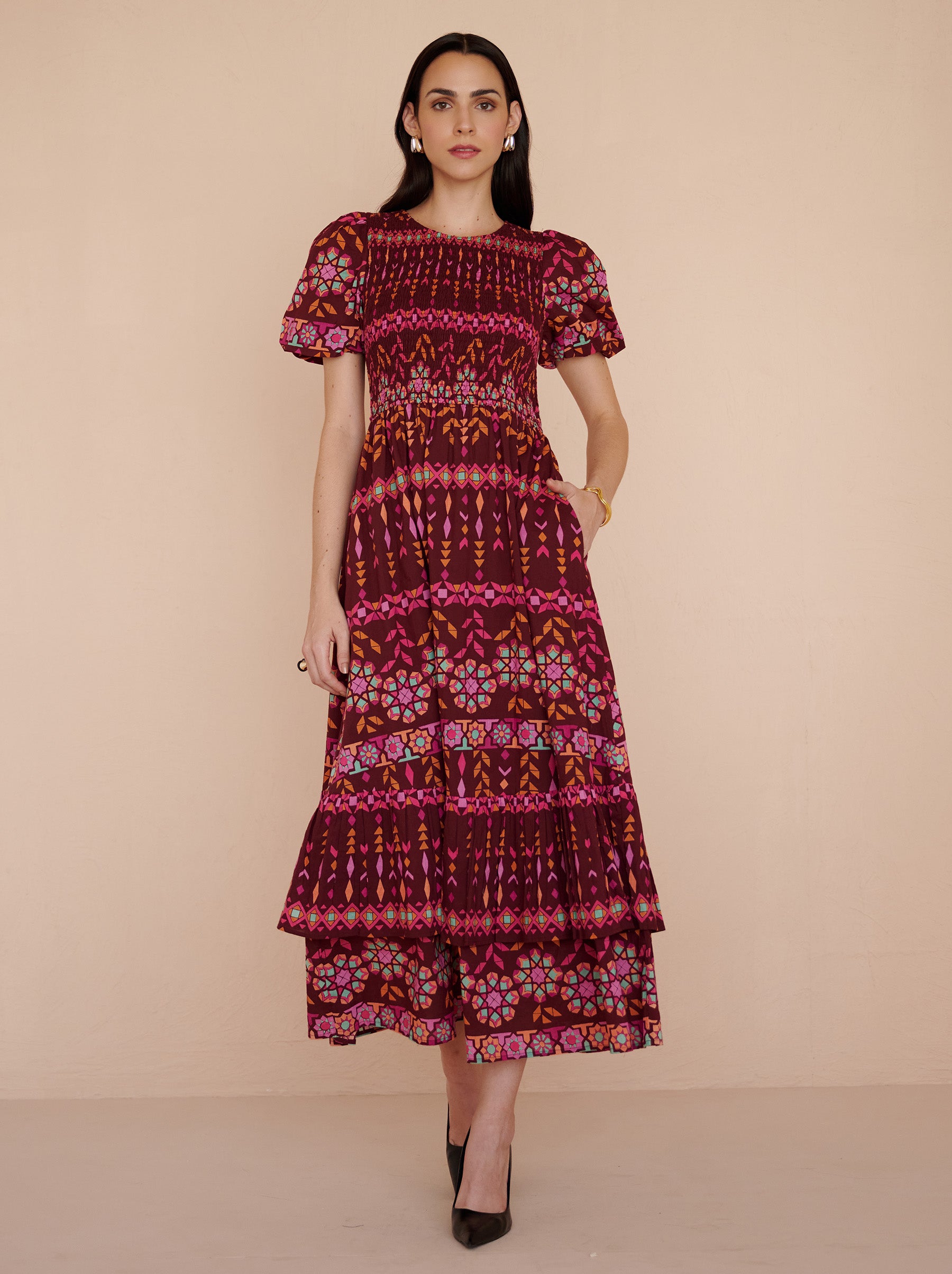 Front view of a model walking with her left hand in pocket. She is wearing the Quant midi dress in the Tapestry print in Rhubarb color. It is featuring a smocked bodice, subtly puffed sleeves, a full tiered skirt, full lining, and side-seam pockets.