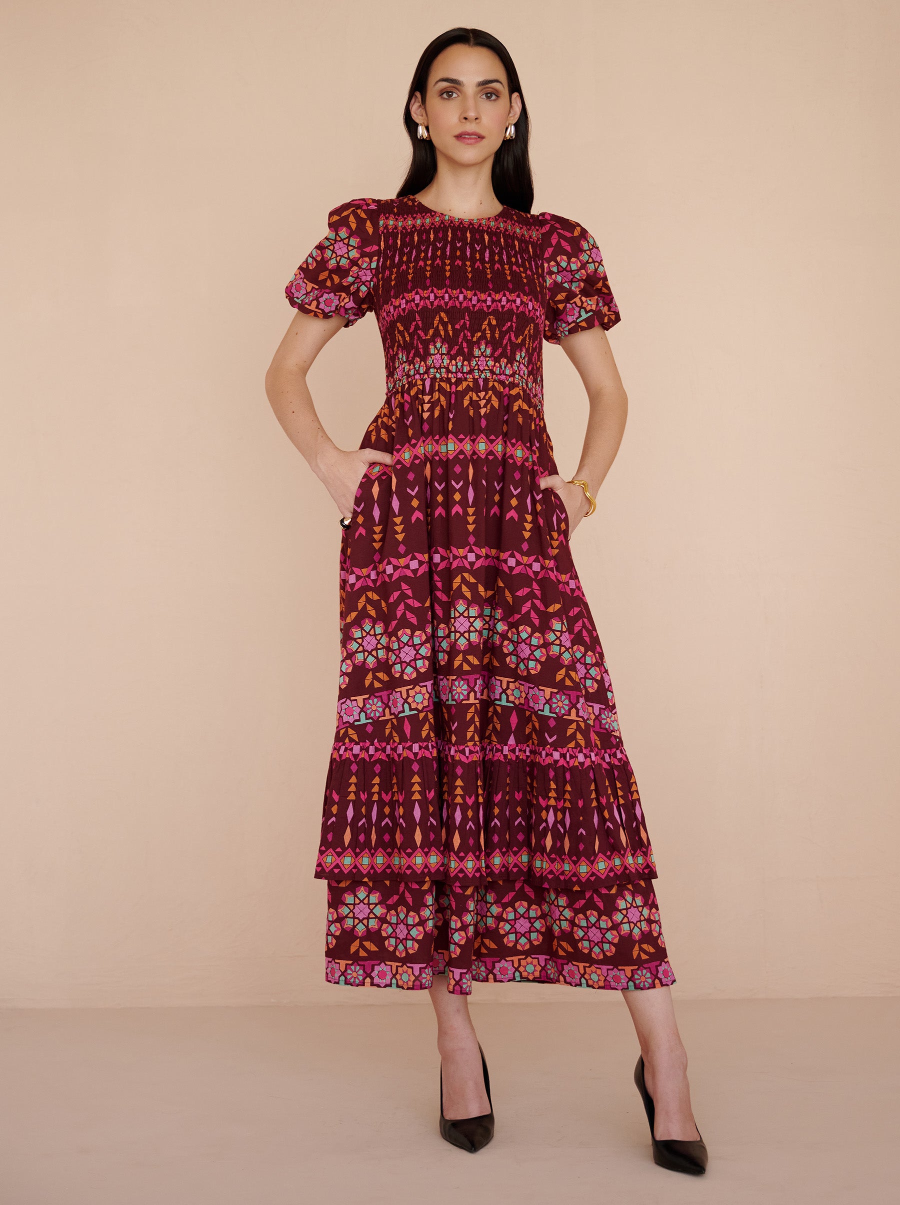 Front view of a model standing with her both hands in pocket. She is wearing the Quant midi dress in the Tapestry print in Rhubarb color. It is featuring a smocked bodice, subtly puffed sleeves, a full tiered skirt, full lining, and side-seam pockets.