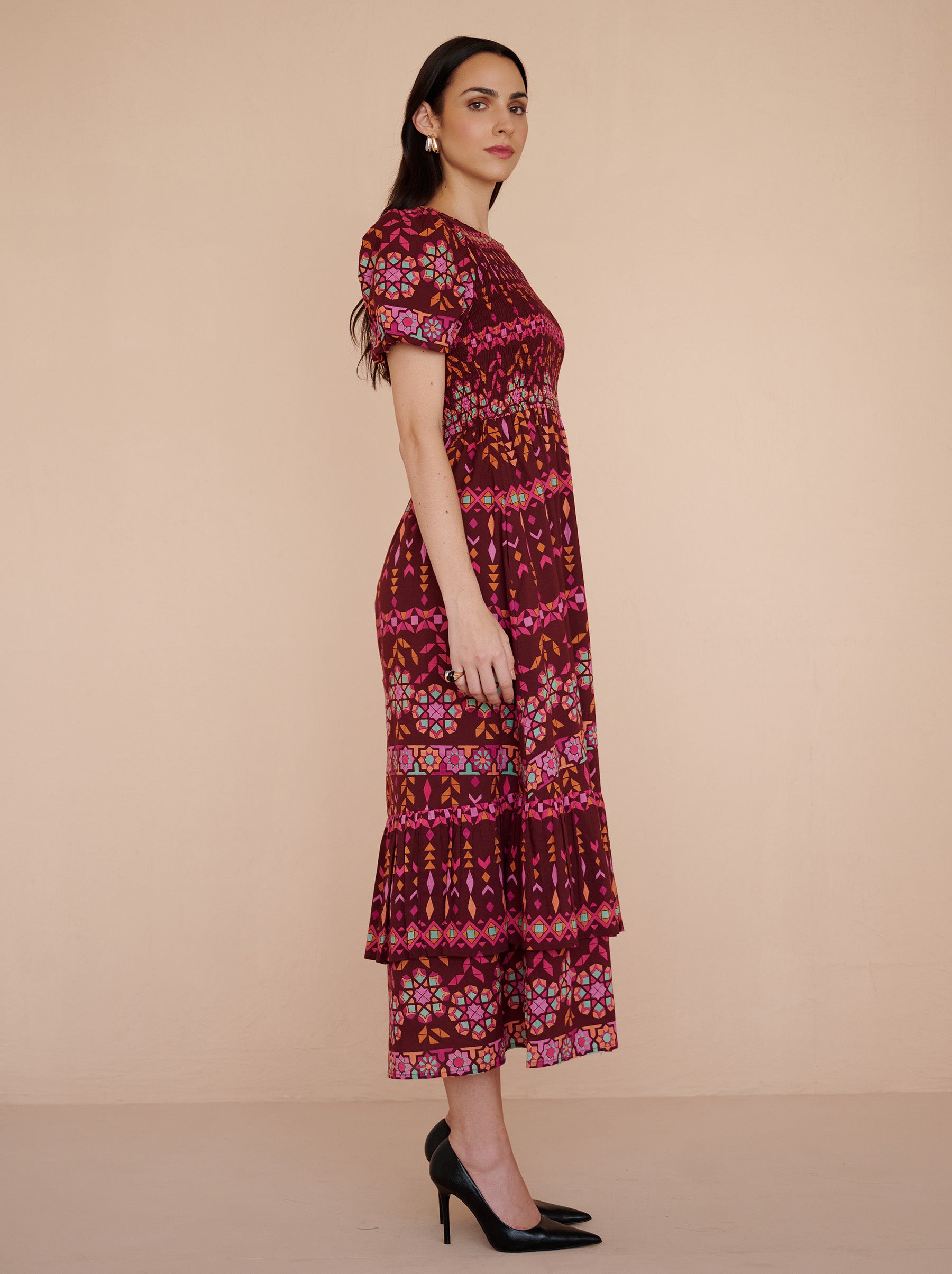 Side view of a model standing. She is wearing the Quant midi dress in the Tapestry print in Rhubarb color. It is featuring a smocked bodice, subtly puffed sleeves, a full tiered skirt, full lining, and side-seam pockets.