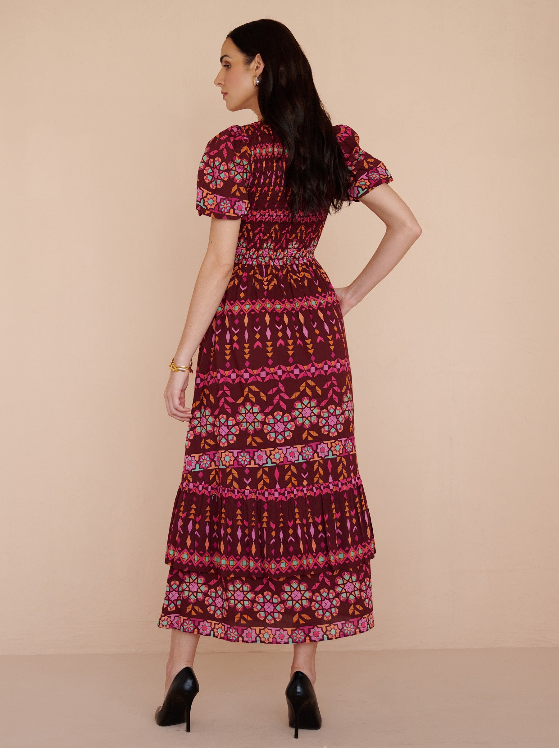 Back view of a model standing. She is wearing the Quant midi dress in the Tapestry print in Rhubarb color. It is featuring a smocked bodice, subtly puffed sleeves, a full tiered skirt, full lining, and side-seam pockets.