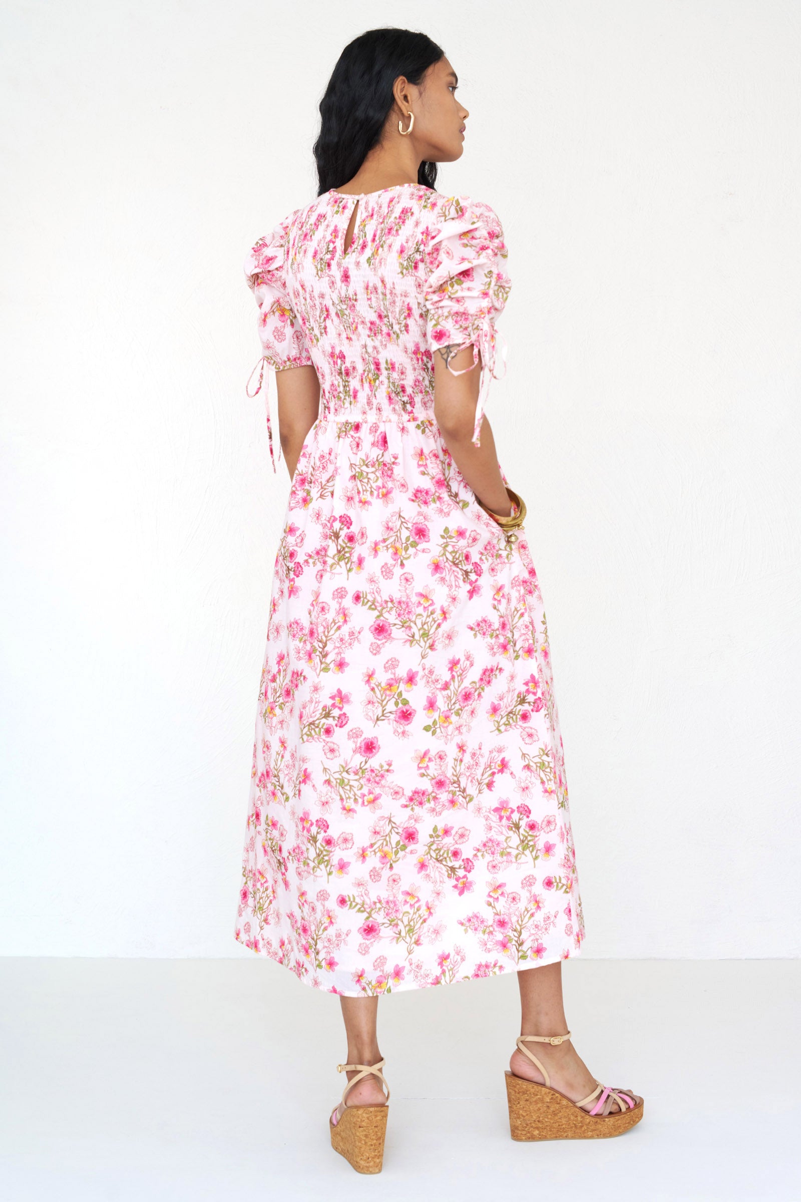Back view of a woman standing wearing the Queenie midi dress in Burdock floral print, featuring a smocked bodice, drawstring sleeves, full skirt & pockets.