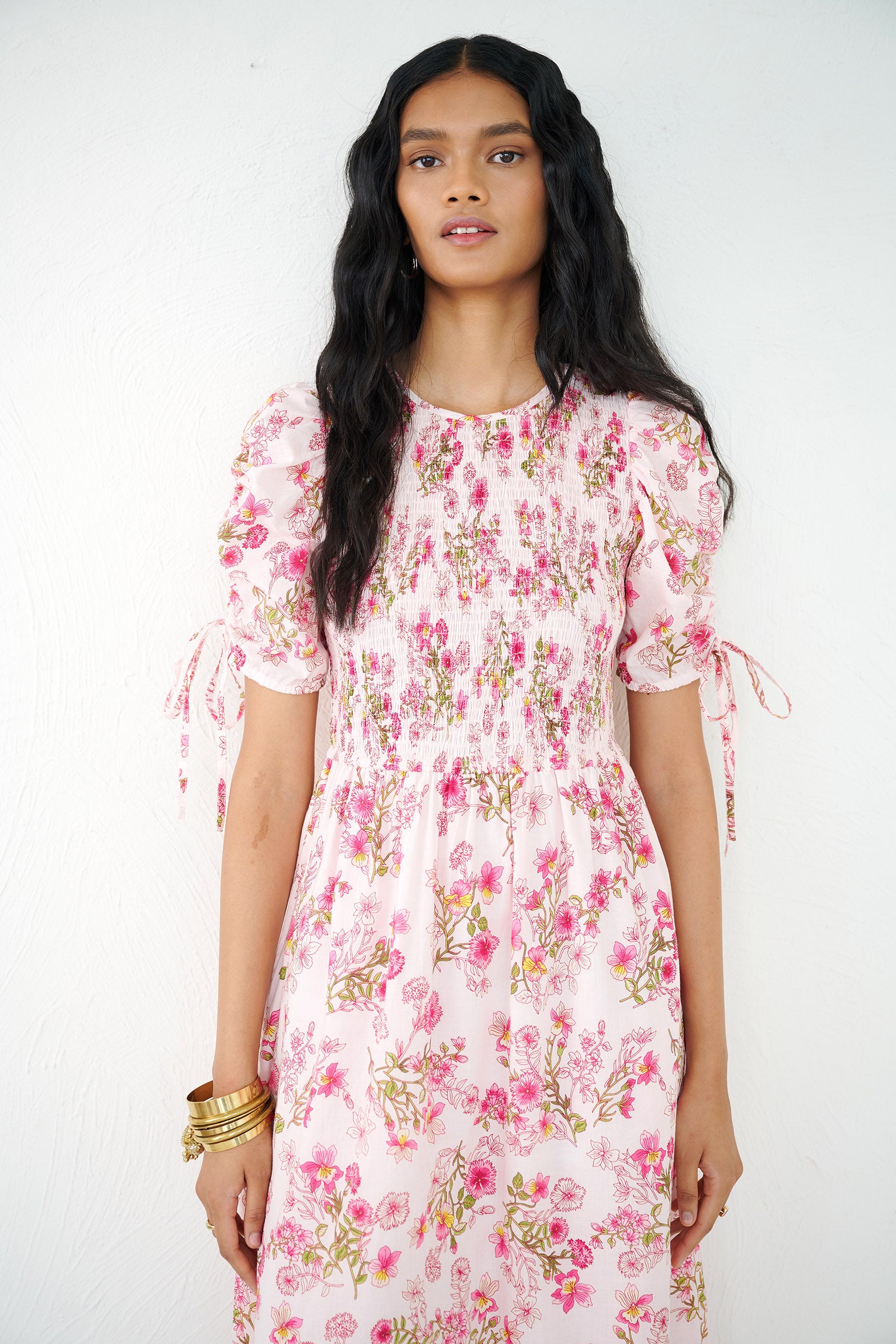 Front view close up of a woman standing wearing the Queenie midi dress in Burdock floral print, featuring a smocked bodice, drawstring sleeves, full skirt & pockets.