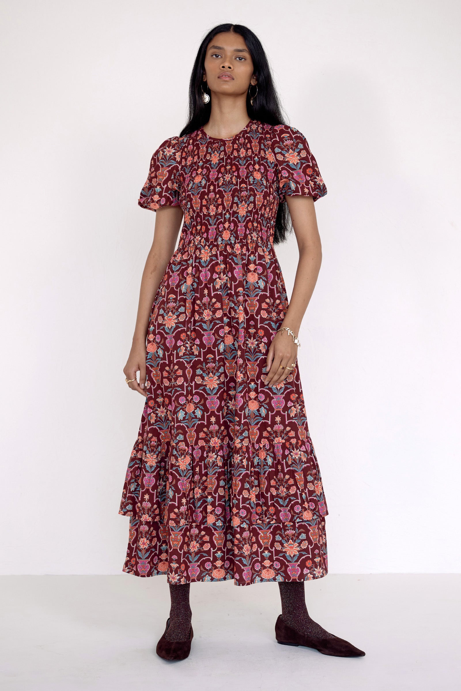 Front view of model standing. She is  wearing the Quant dress in Floral Vase print. It features a smocked bodice, puffed sleeves, and a full tiered skirt with pockets.