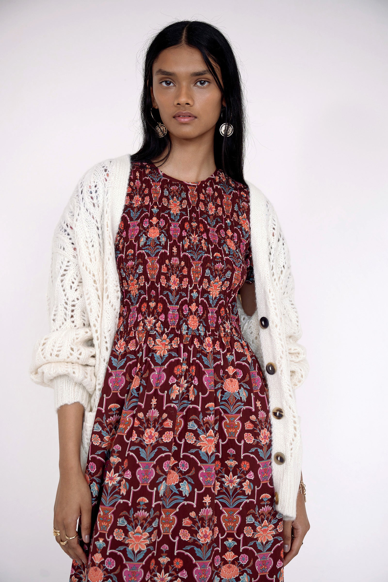 Close up of front view of model standing. She is  wearing the Quant dress in Floral Vase print. It features a smocked bodice, puffed sleeves, and a full tiered skirt with pockets. Styled with an ivory cardigan.