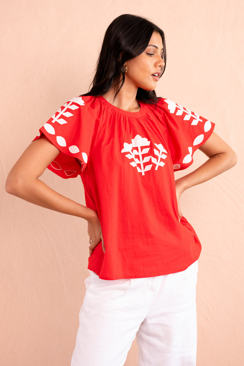 Front view of woman wearing a red short-sleeve top with white appliqué embroidery in geometric floral motifs on the chest and sleeves, paired with white pants.