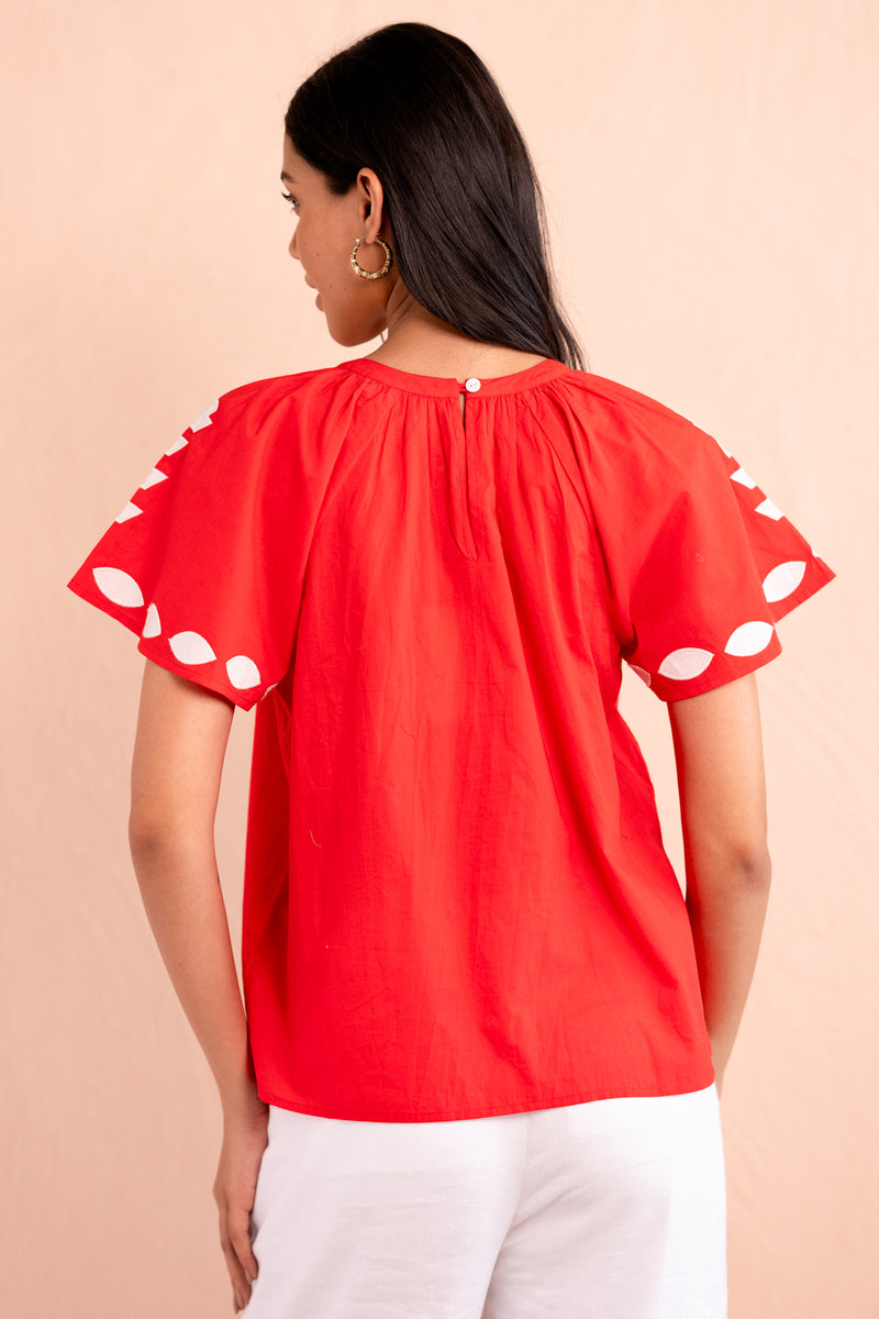 Back view of woman wearing a red short-sleeve top with white appliqué embroidery in geometric floral motifs on the chest and sleeves, paired with white pants.
