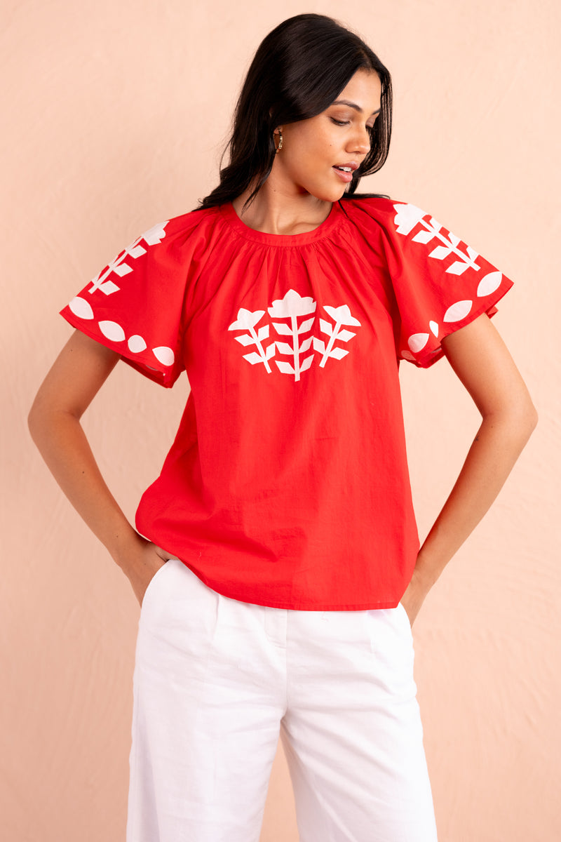 Front view of woman wearing a red short-sleeve top with white appliqué embroidery in geometric floral motifs on the chest and sleeves, paired with white pants.