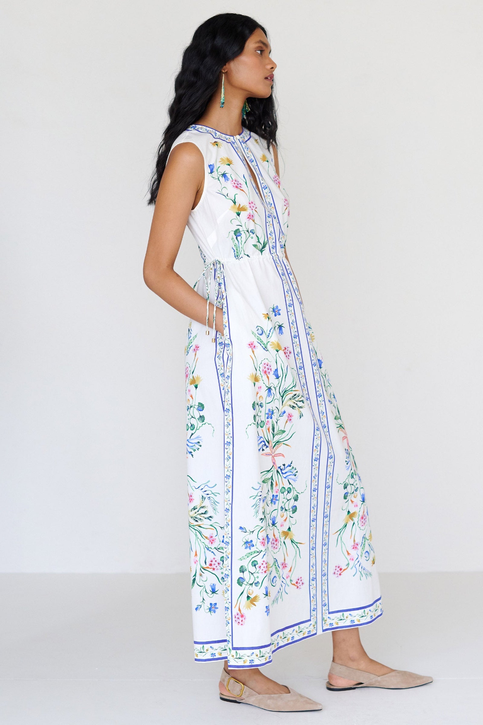 Side view close up of a woman standing with hands in pocket. She is wearing the Rhonda column dress in Tidepool print. It features a crewneck with a fabric-covered button and keyhole detail, side drawstrings with gold caps, and side-seam pockets.