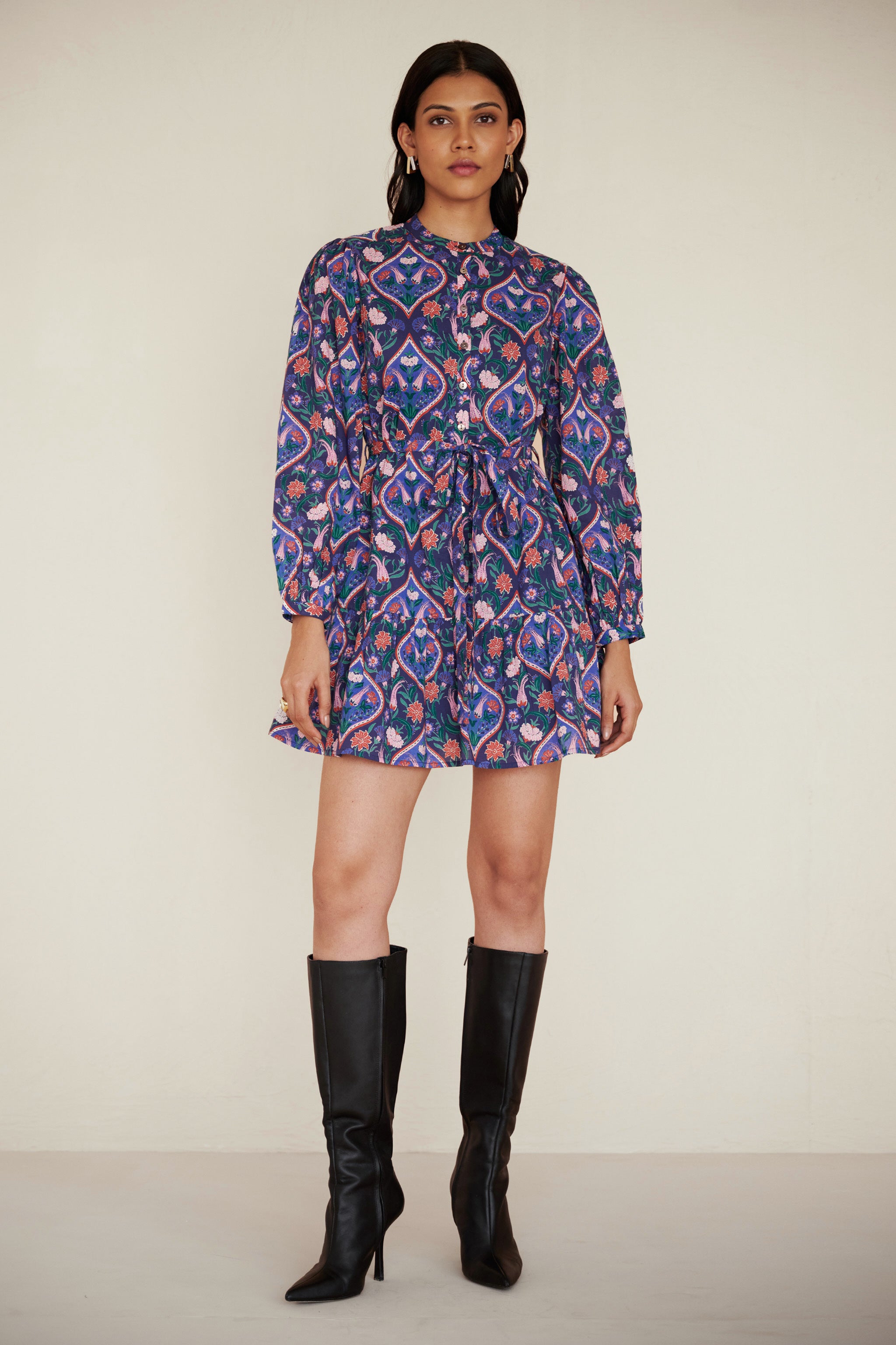 Woman standing straight wearing Roma dress in Persian Tile Lapis print. Its a cotton mini dress with wrist-length sleeves, a floral tile print, and a detachable belt, styled with side pockets.