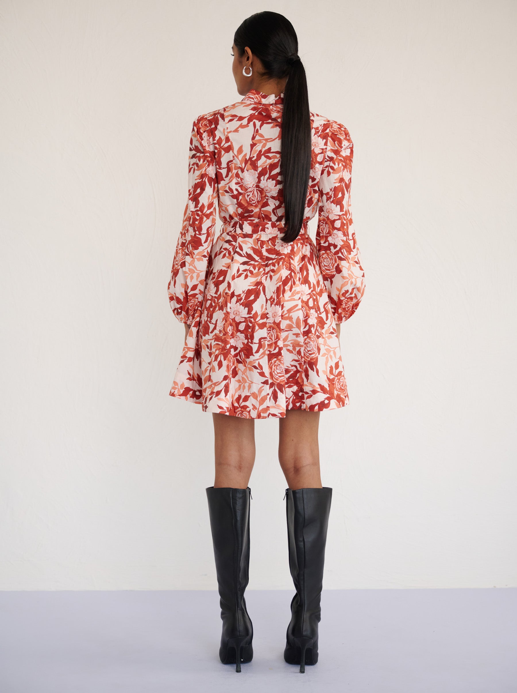 Back view of a model standing. She is wearing the Rowena mini shirtdress in crisp cotton poplin in Bibi Rose Chocolate print. It is featuring a delicate collar, lantern sleeves, short hem, full lining, and side-seam pockets.