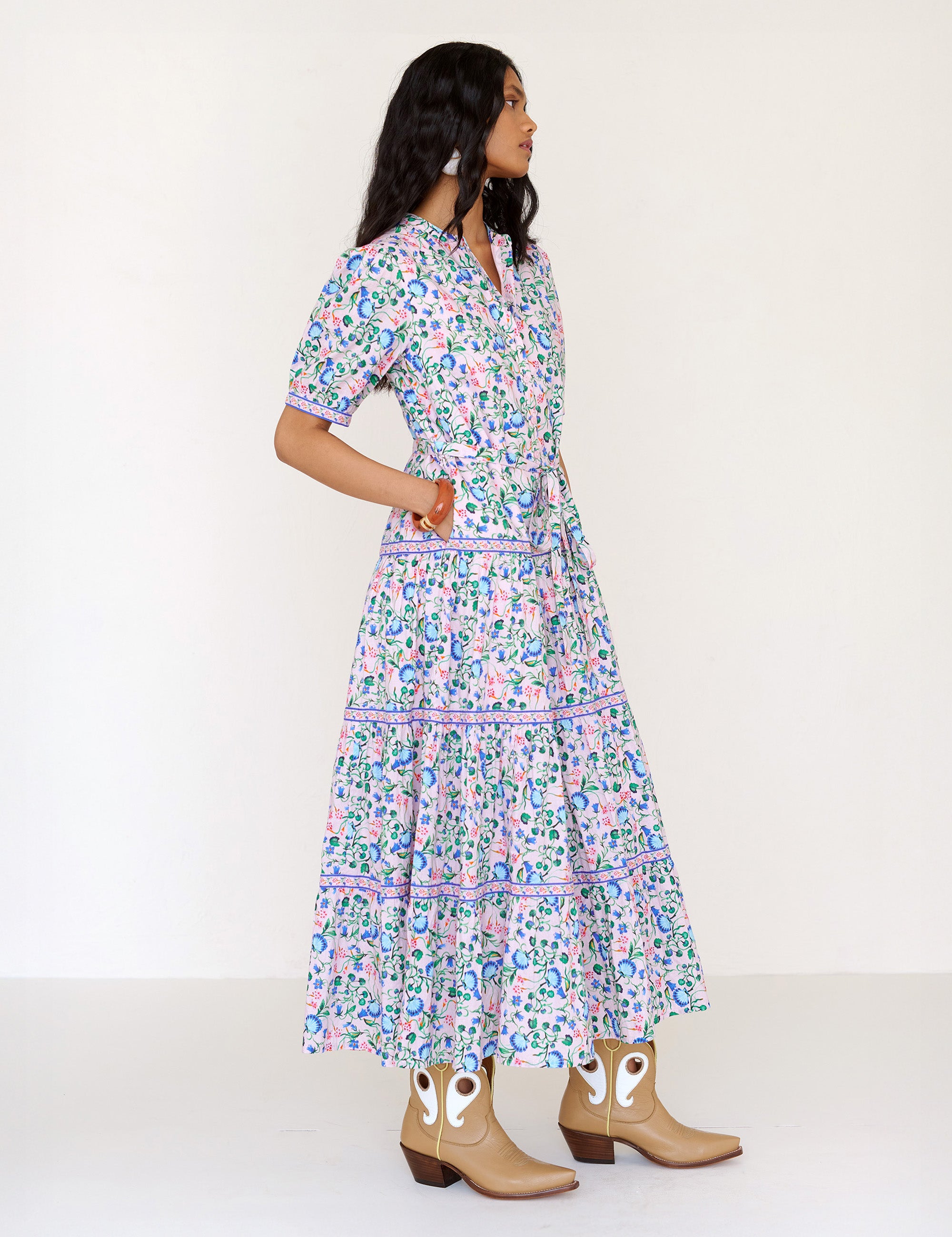 Side view of a woman standing. She is  wearing the Saylor dress in Clam Garland floral print with her hands in pocket. It features a classic shirtdress design with stand collar, tiered skirt, contrast trim, and a detachable belt.