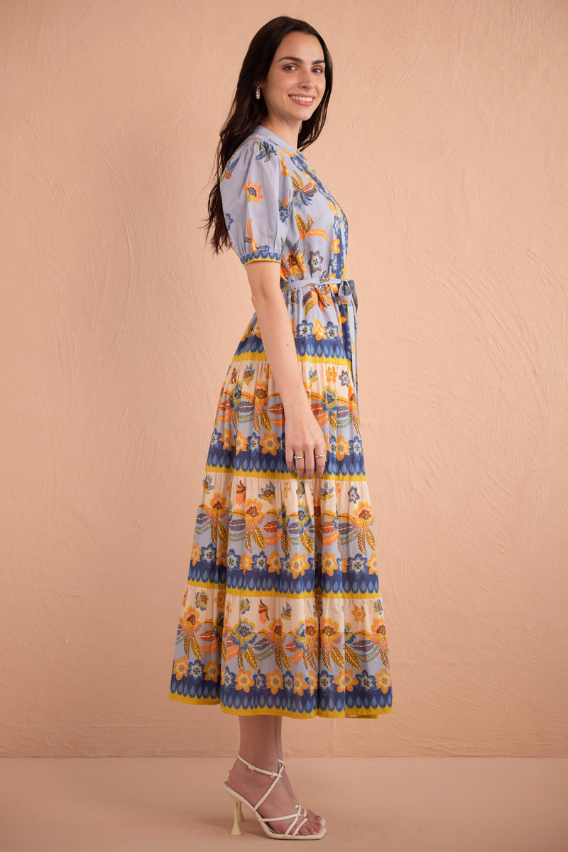Side view of woman wearing floral print midi dress with short puff sleeves, tiered skirt, and tie waist in amber, blue, and yellow tones.