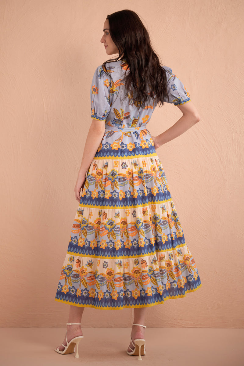 Back view of woman wearing floral print midi dress with short puff sleeves, tiered skirt, and tie waist in amber, blue, and yellow tones.