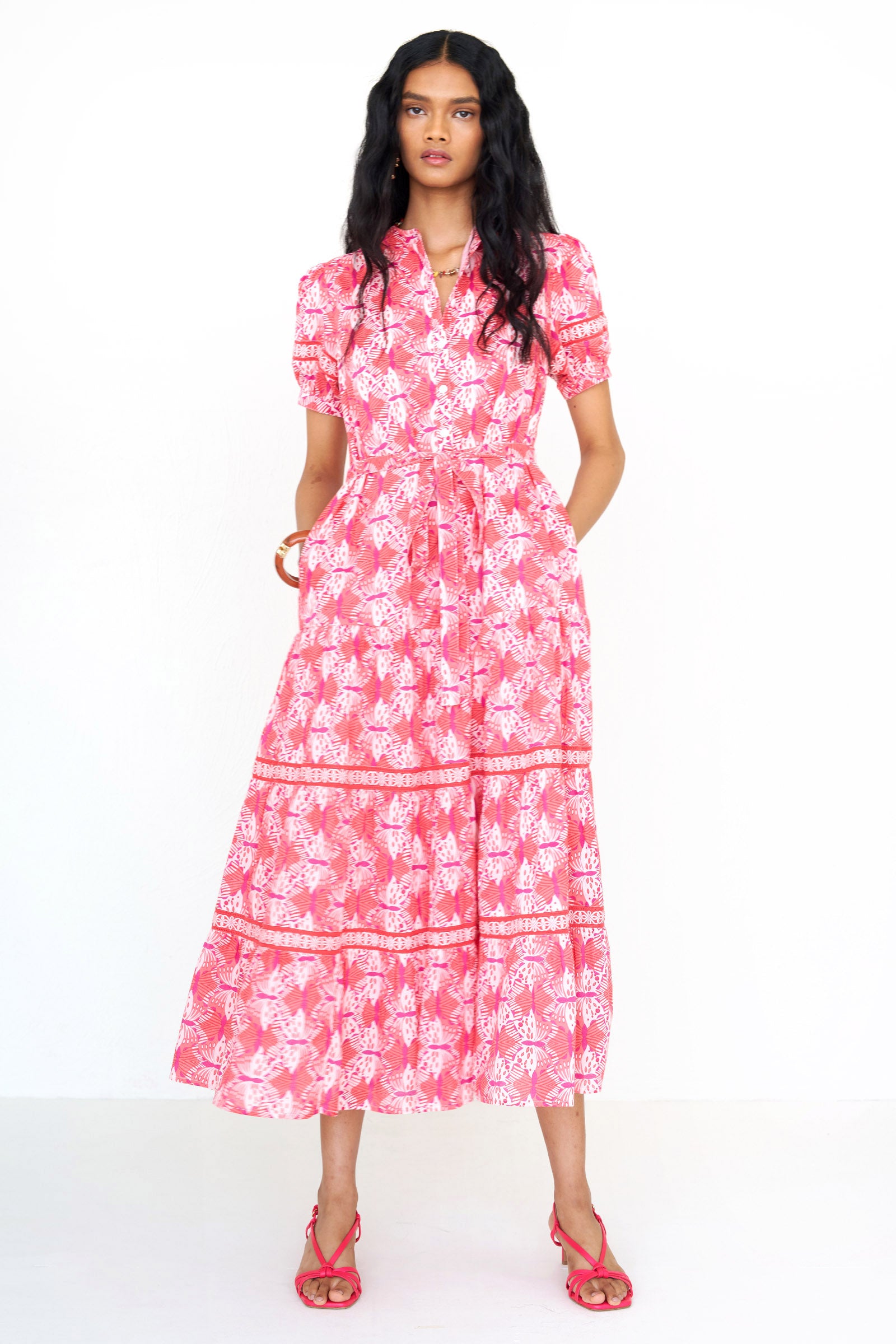 Front view of a woman standing wearing the Saylor dress in Flutter print with her hands in pockets. It features a stand collar, tiered skirt, detachable belt, and celtic-knot ladder lace detailing in a full-length shirtdress silhouette.