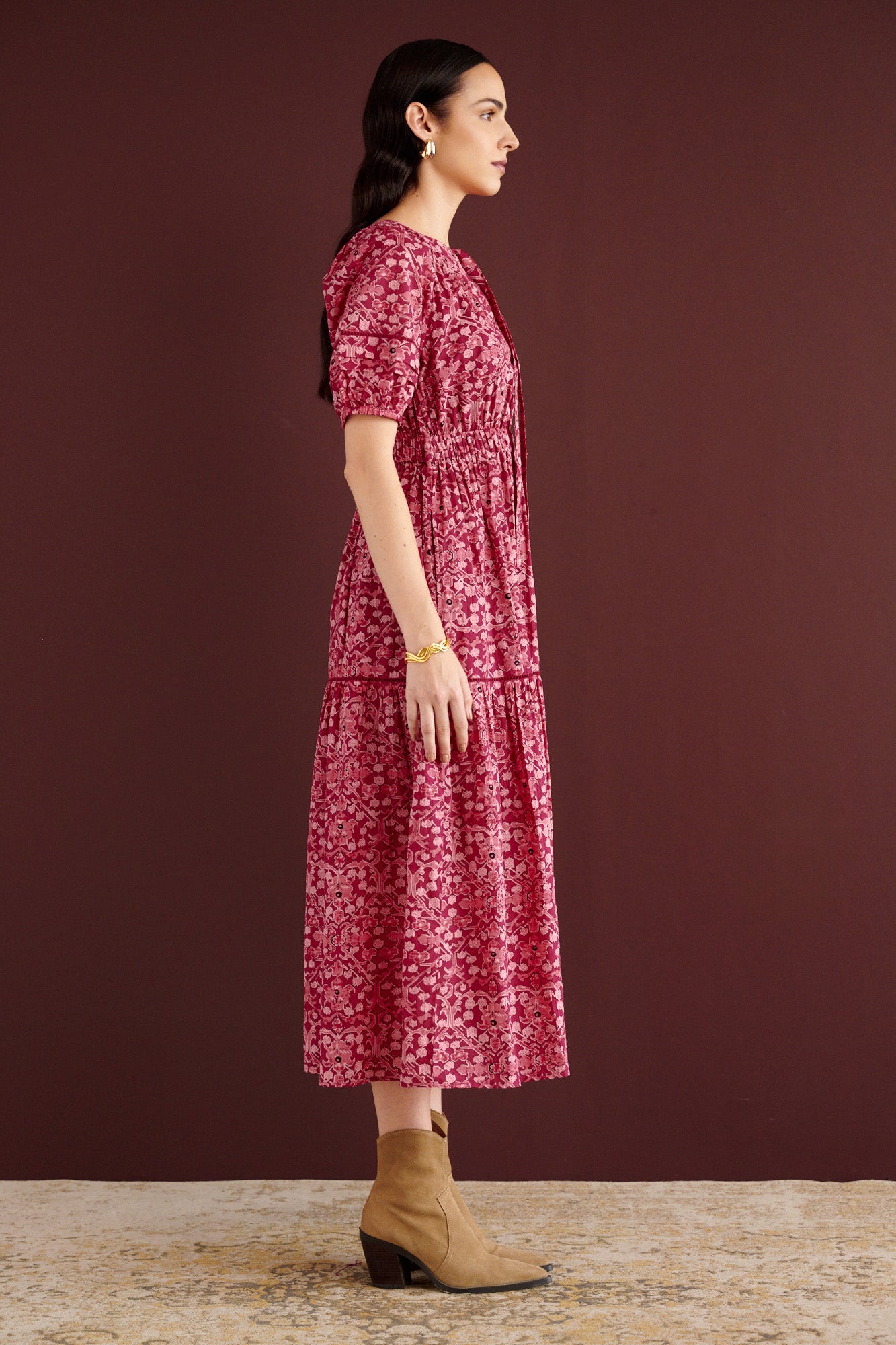 Side view of woman standing. She is wearing the Selma full-length V-neck dress in Ottoman print, with tie-neck, elastic waist, lace insets at skirt and sleeves, smooth cotton cambric, side-seam pockets, and fully lined construction.