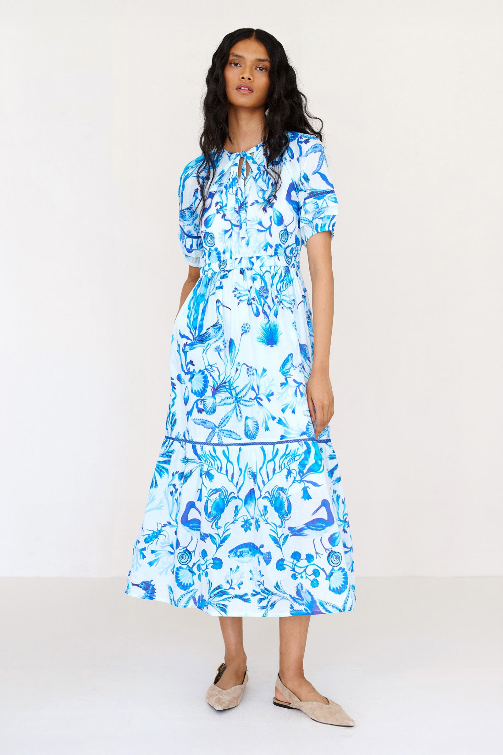 Front view of a woman standing. She is wearing the Selma full-length V-neck dress in tonal Tidepool print. It features a tie-neck, elasticated waist, ladder lace detailing, and side-seam pockets.