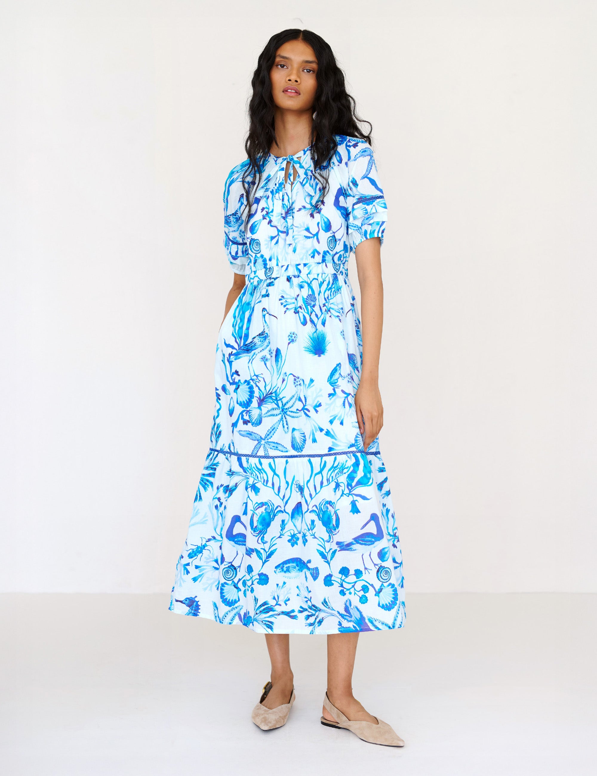 Front view of a woman standing. She is wearing the Selma full-length V-neck dress in tonal Tidepool print. It features a tie-neck, elasticated waist, ladder lace detailing, and side-seam pockets.