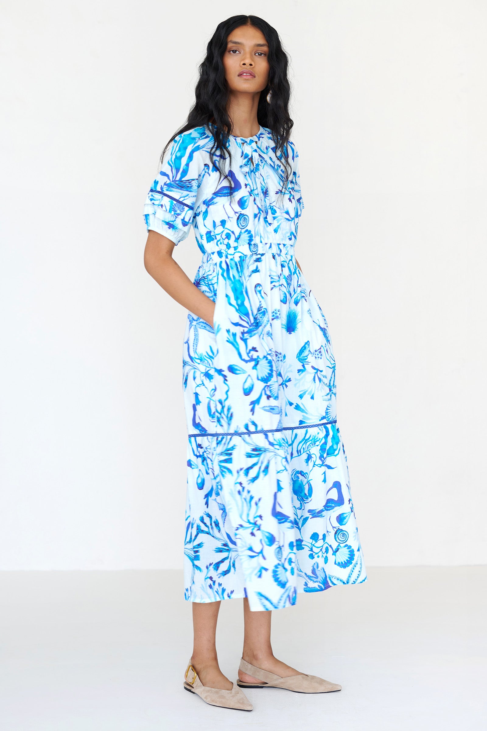 Side view of a woman standing wearing the Selma dress in Tidepool print, featuring a V-neck with tie, elastic waist, ladder lace insets, full-length skirt and side-seam pockets.