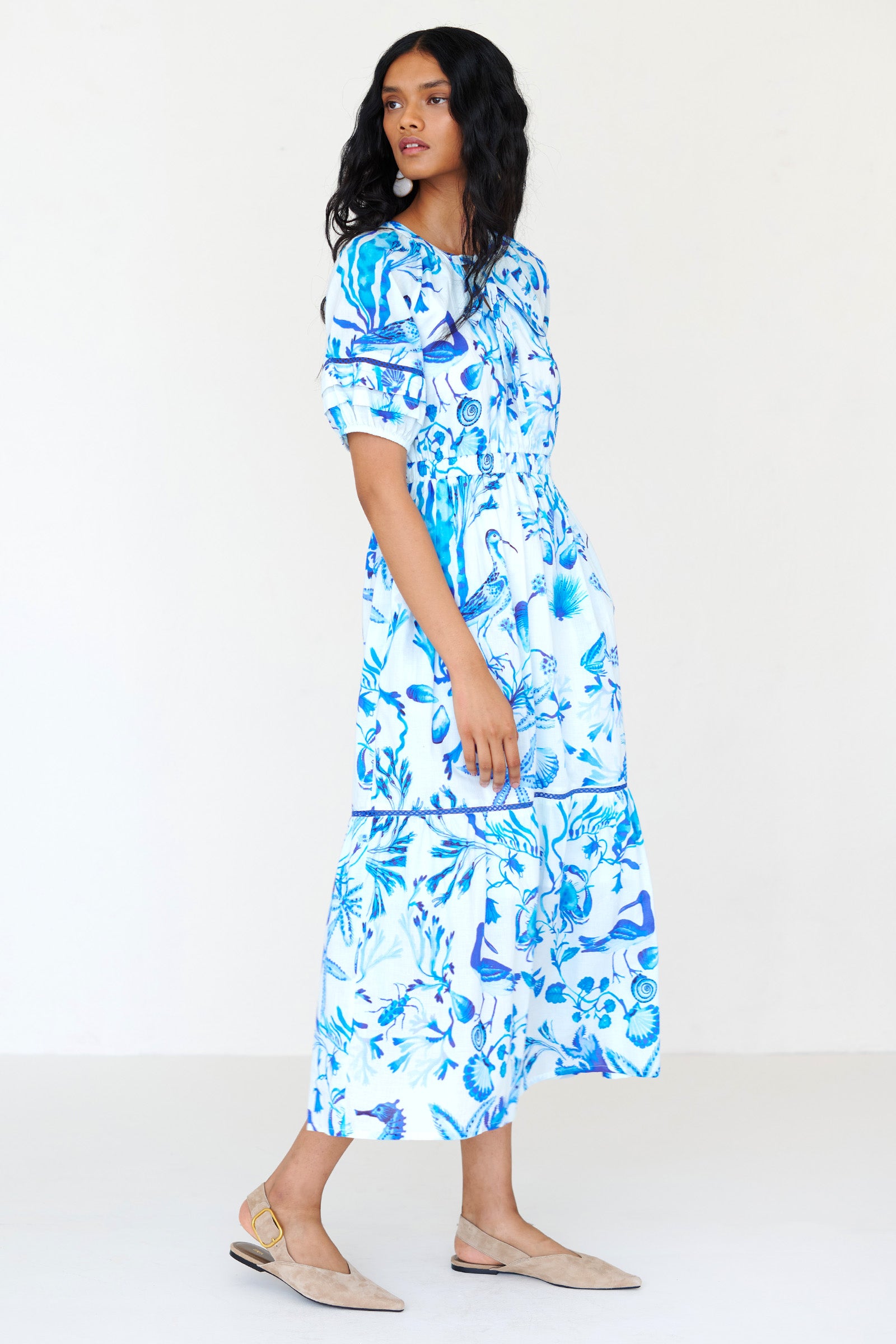 Side view of a woman standing wearing the Selma dress in Tidepool print, featuring a V-neck with tie, elastic waist, ladder lace insets, full-length skirt and side-seam pockets.