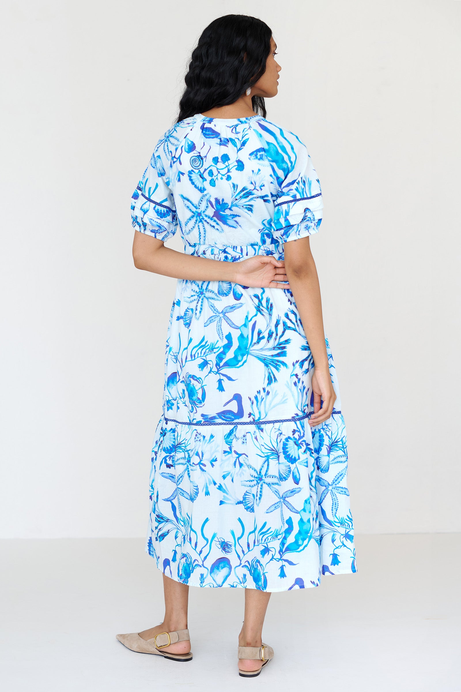 Back view of a woman standing wearing the Selma dress in Tidepool print, featuring a V-neck with tie, elastic waist, ladder lace insets, full-length skirt and side-seam pockets.